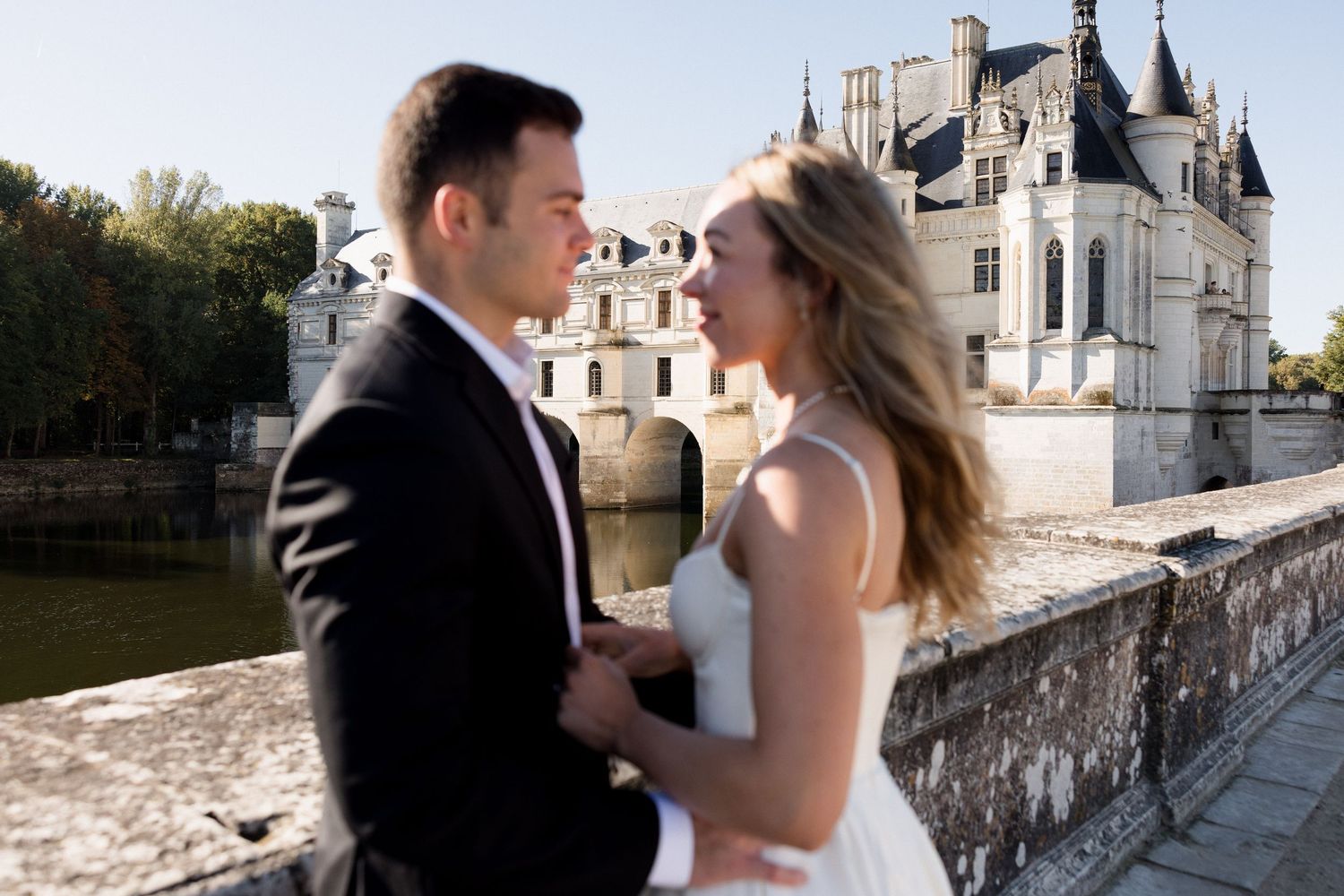 Couple embraces on stone bridge with elegant French chateau reflected in water behind them.