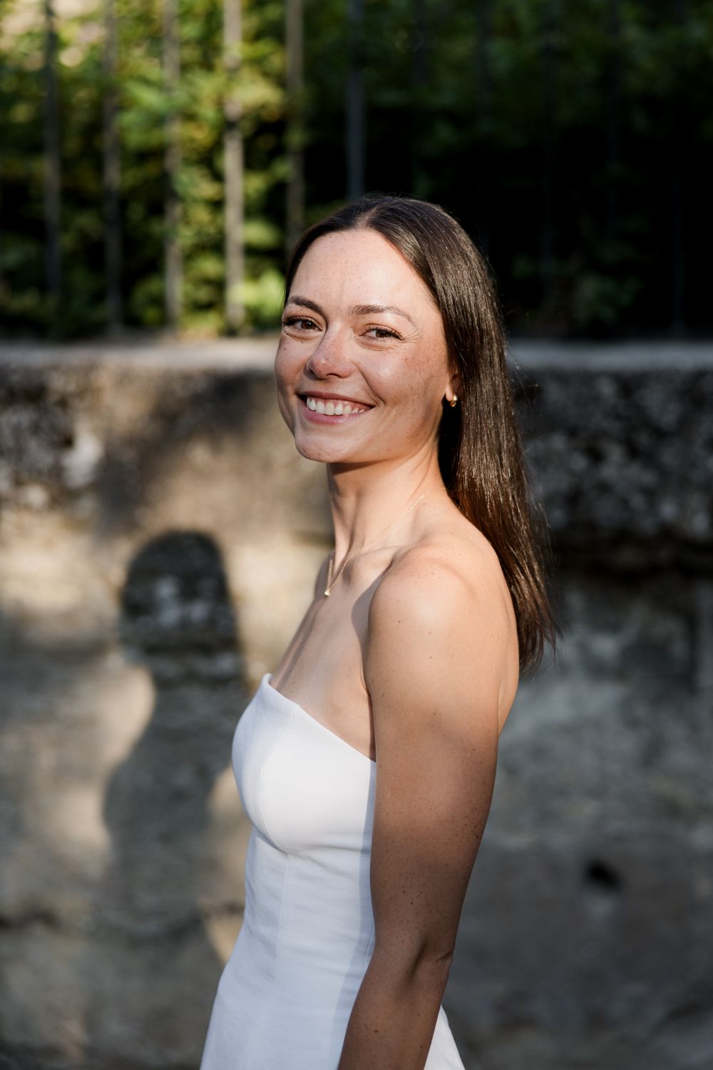 A portrait of someone in a white dress with long brown hair standing against a stone wall outdoors.