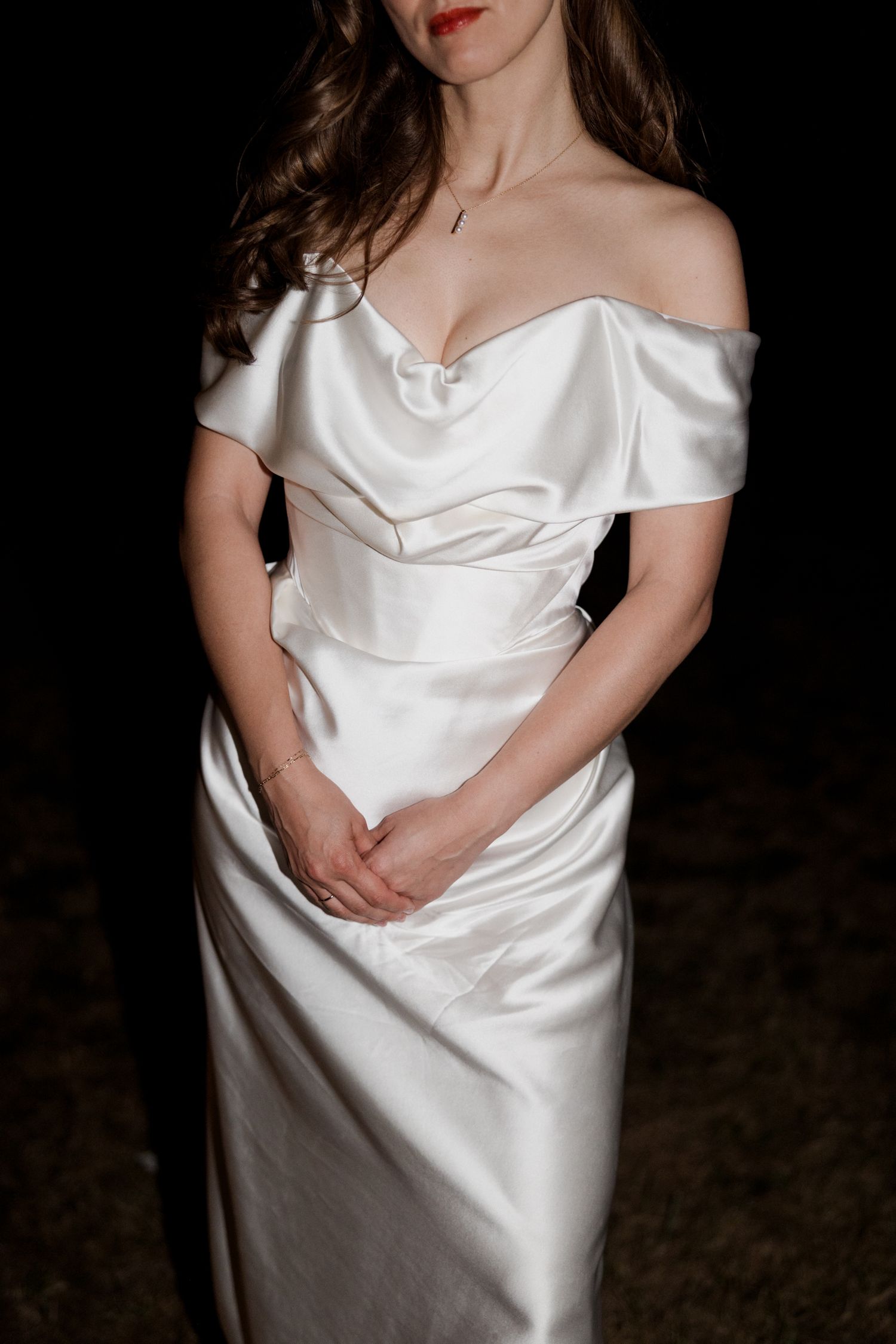 Elegant white satin off-shoulder wedding dress with draped bodice detail shown against dark background.