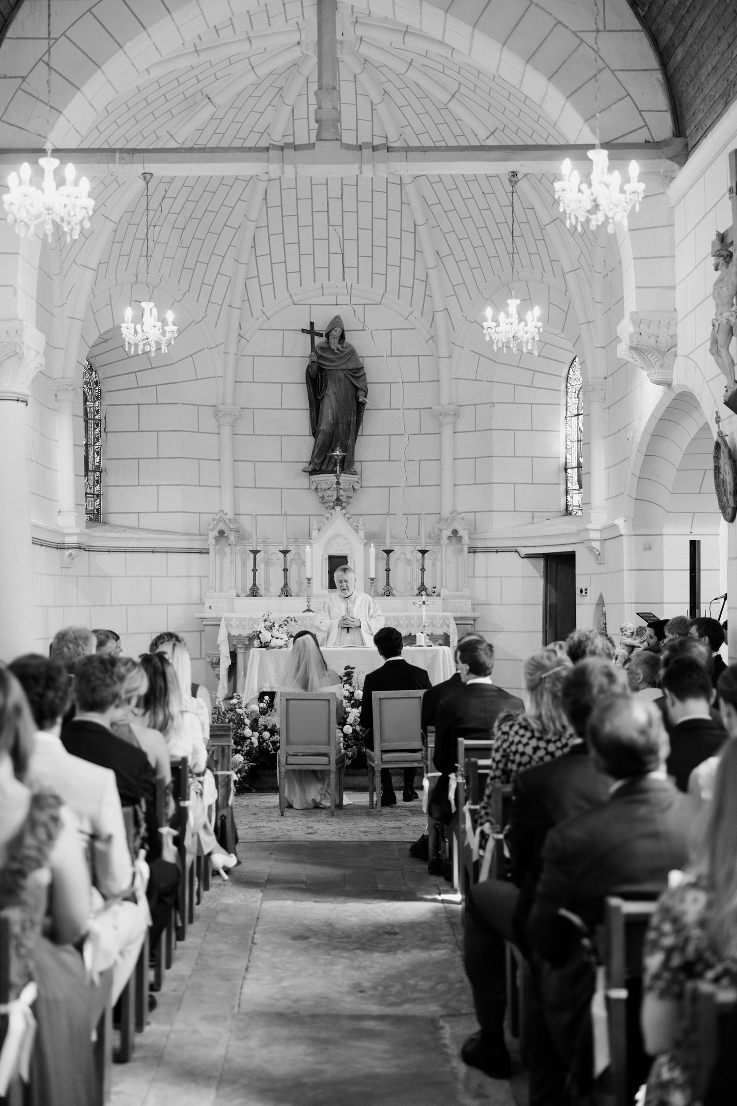 Black and white photo of a church wedding ceremony with elegant chandeliers and guests seated in wooden pews.