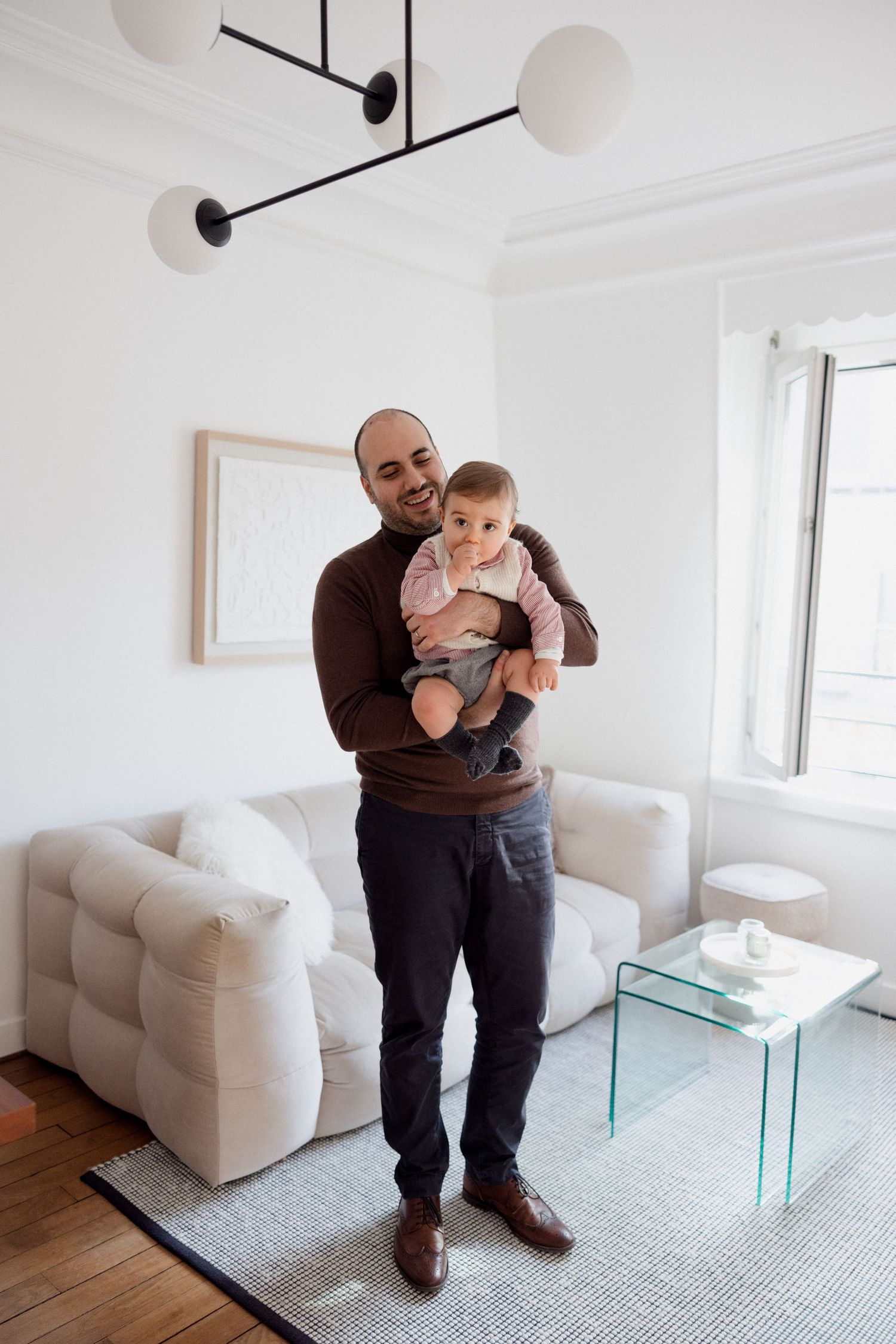 A smiling man holds a baby in a bright, modern living room with white sofa, glass table, and globe pendant light.
