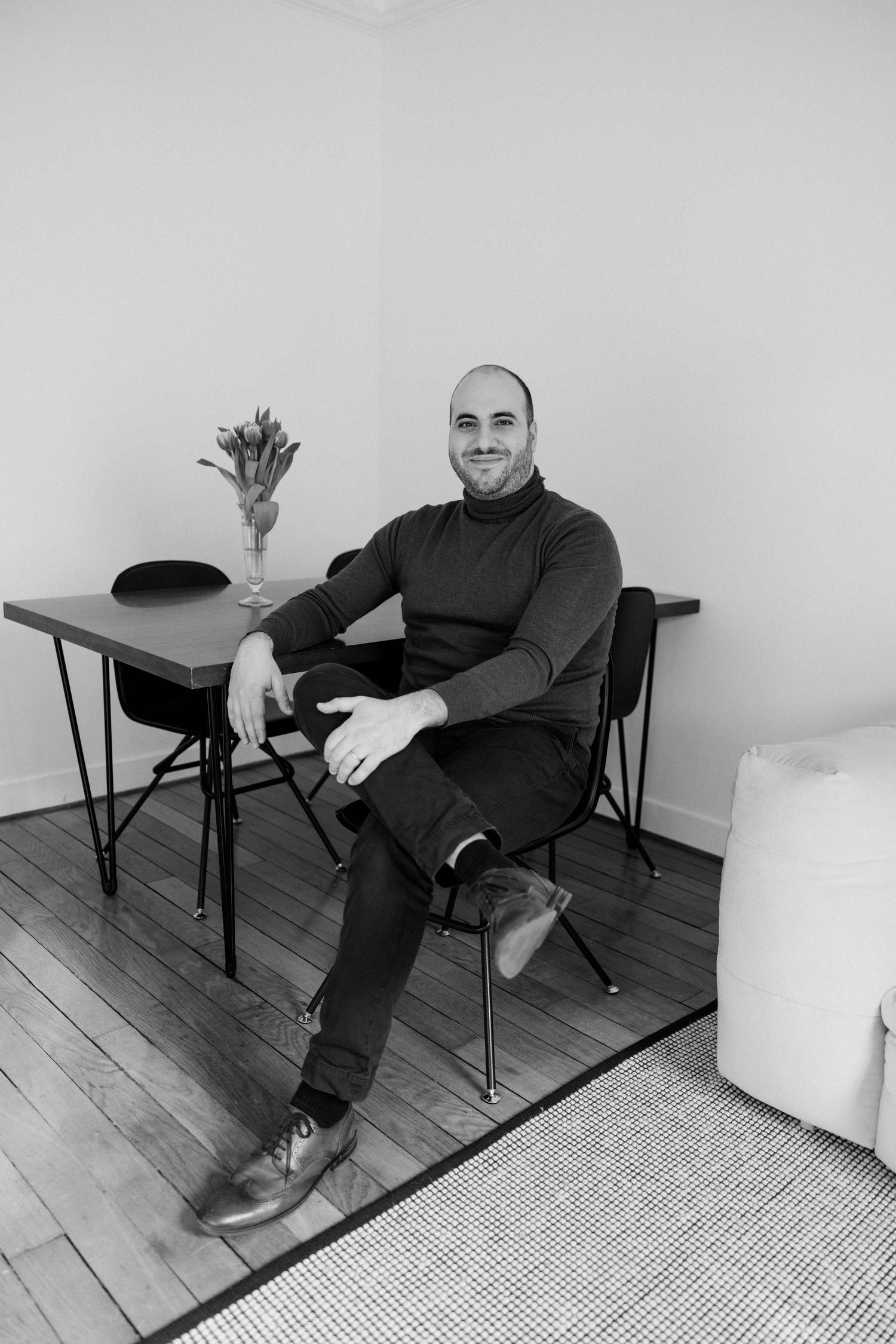 Smiling bald man in turtleneck sweater sitting relaxed on chair at minimalist table in black and white photo.