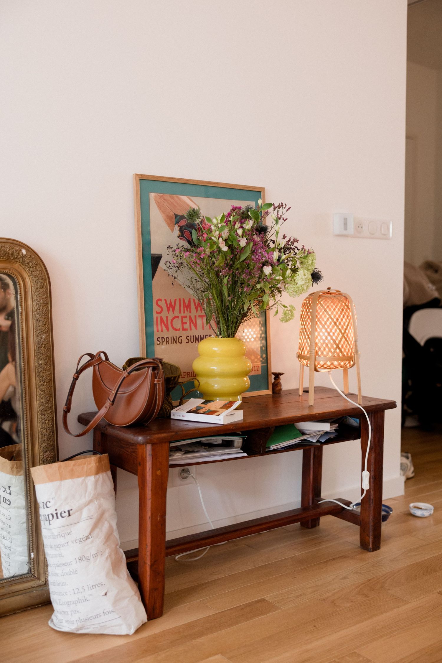 Wooden console table with yellow vase, wildflowers, wicker lamp, framed poster, brown leather bag, and paper sack on hardwood floor.
