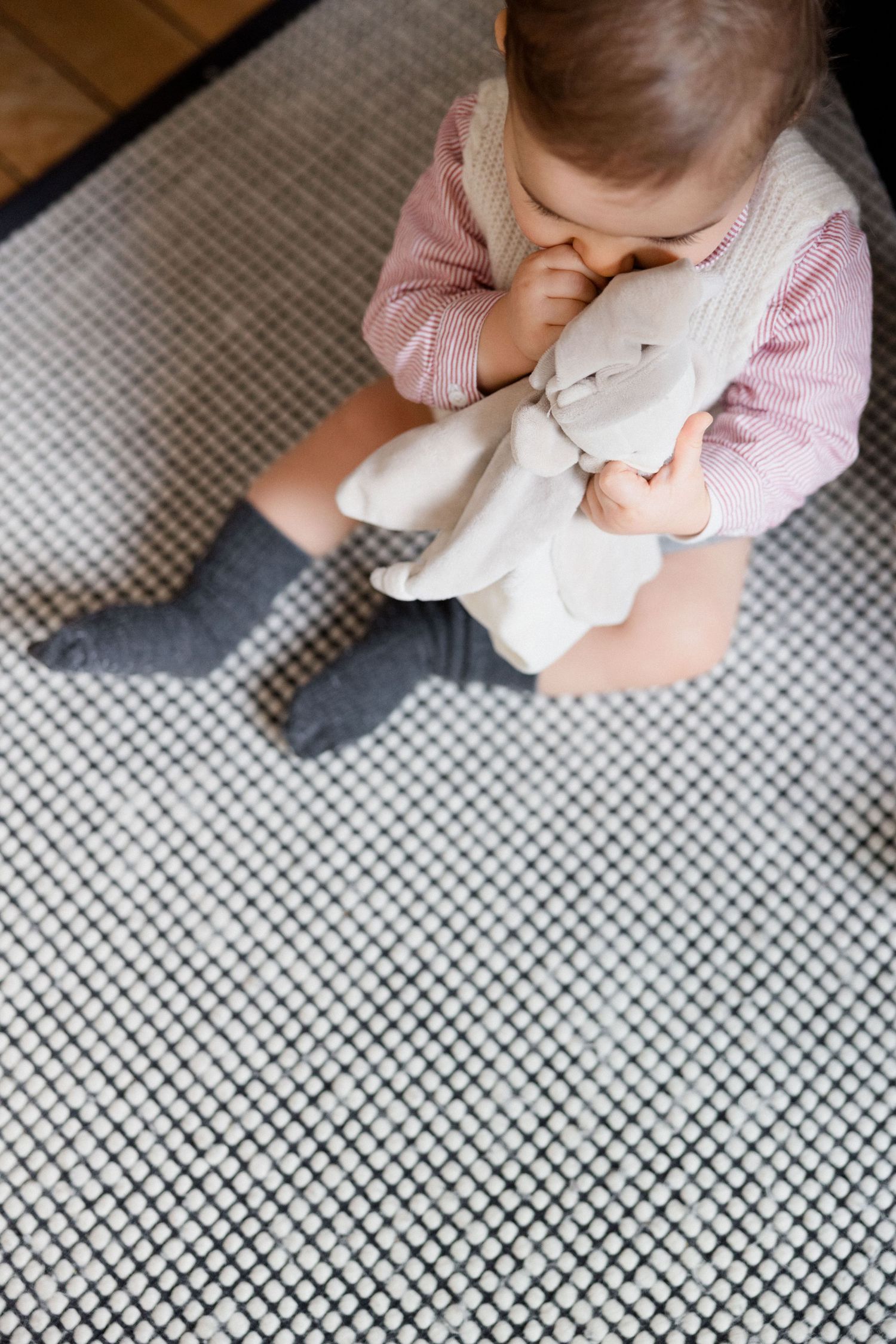 Toddler girl in pink outfit sitting on gray rug, hugging a white stuffed bunny toy while wearing gray socks.