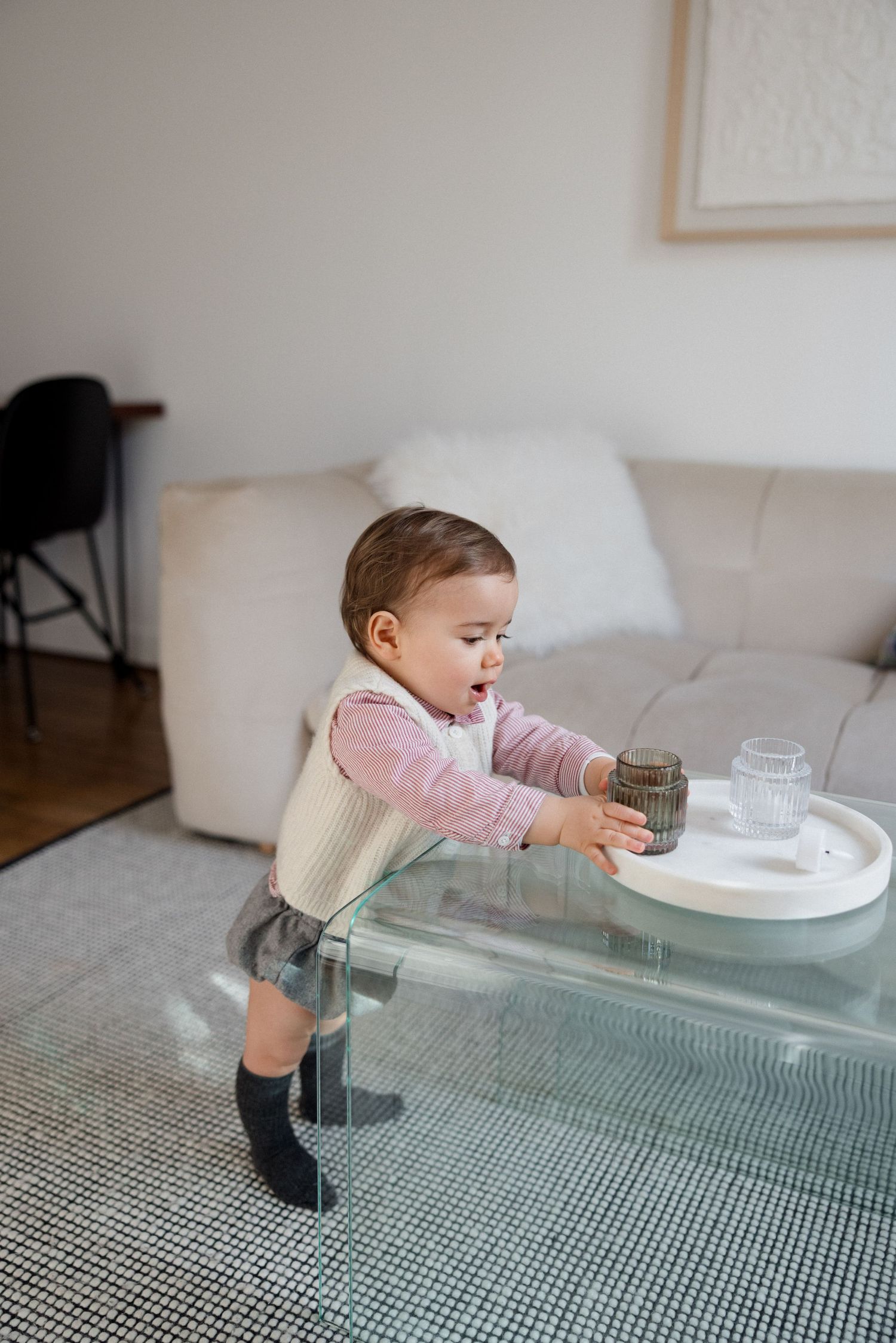 Curious baby in pink sweater and gray skirt reaching for a glass on a modern glass coffee table in a living room.