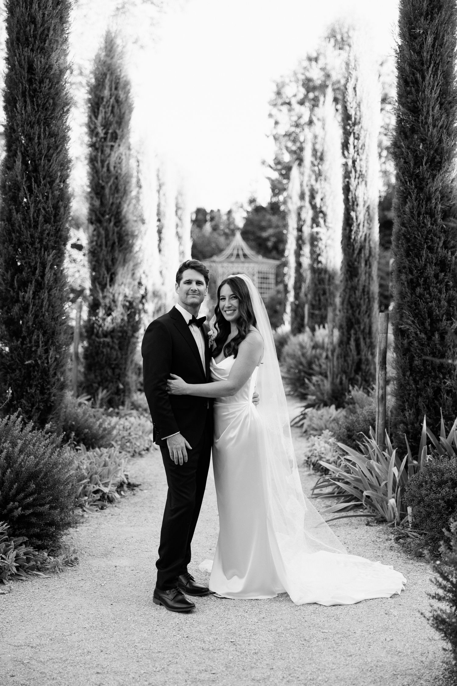 A wedding couple poses on a path lined with tall cypress trees in an elegant black and white portrait. wedding photographer france
