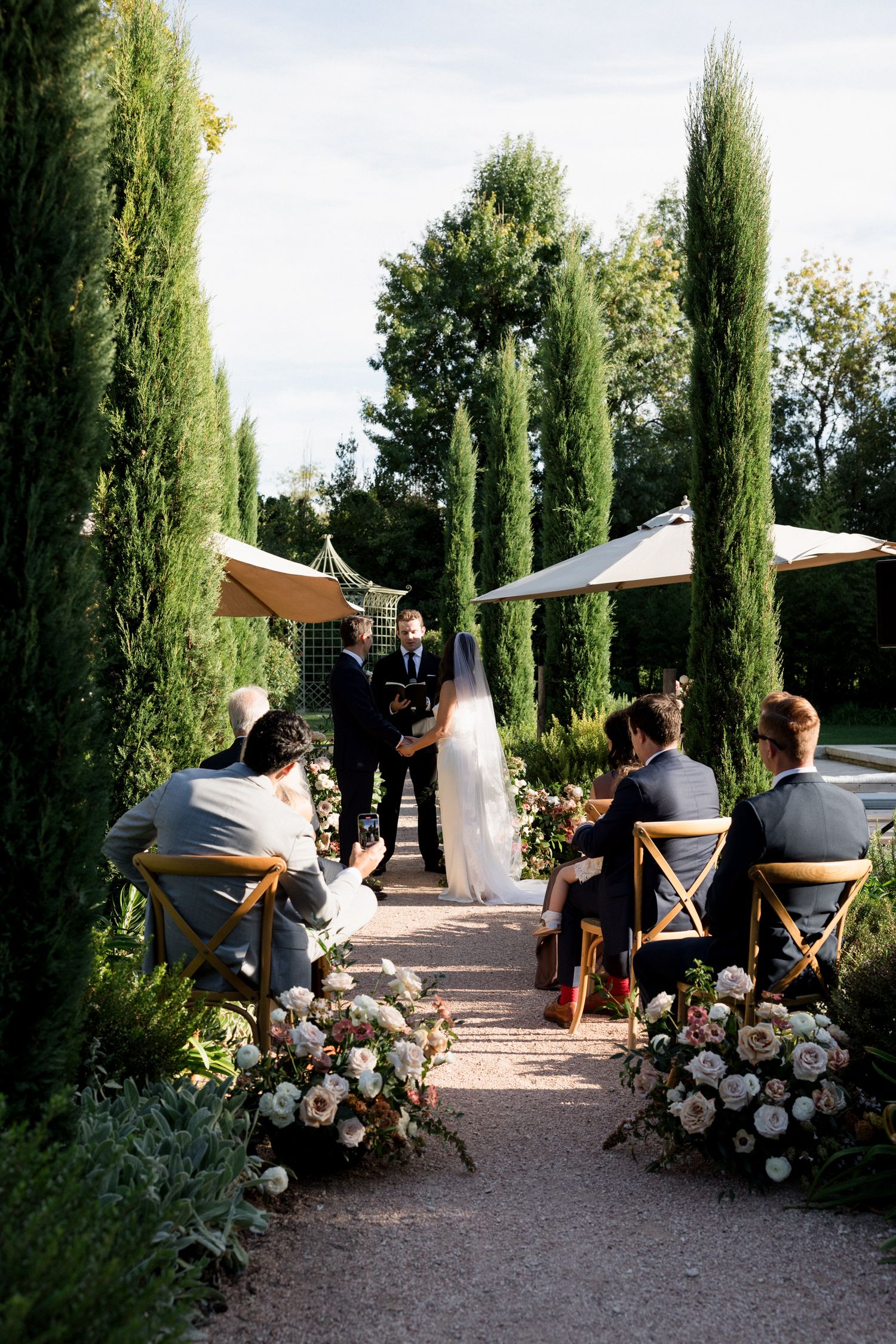 Wedding ceremony setup with tall cypress trees lining a garden path and white floral arrangements.