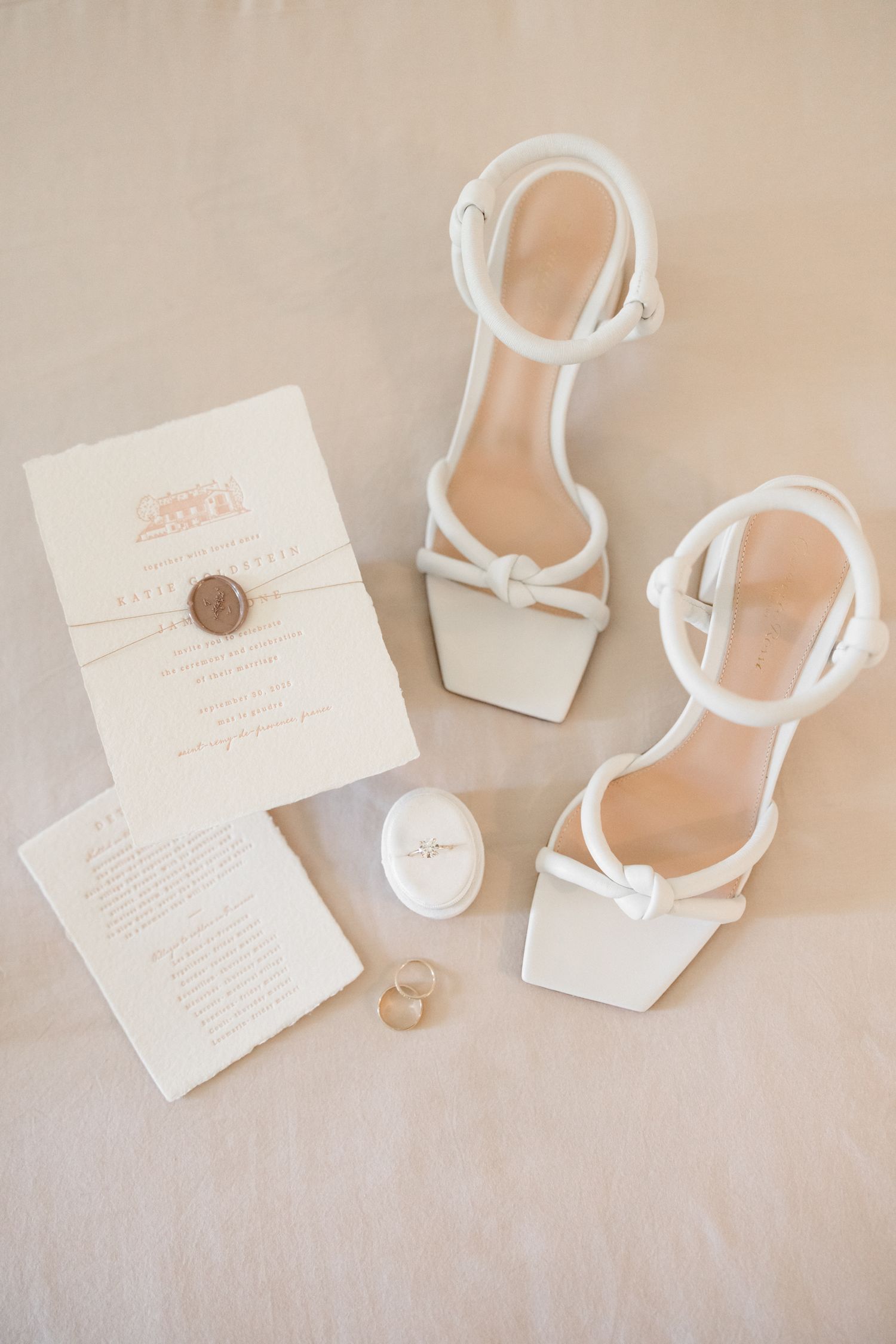 Elegant white bridal accessories including strappy sandals, wedding invitation suite and rings on neutral background.