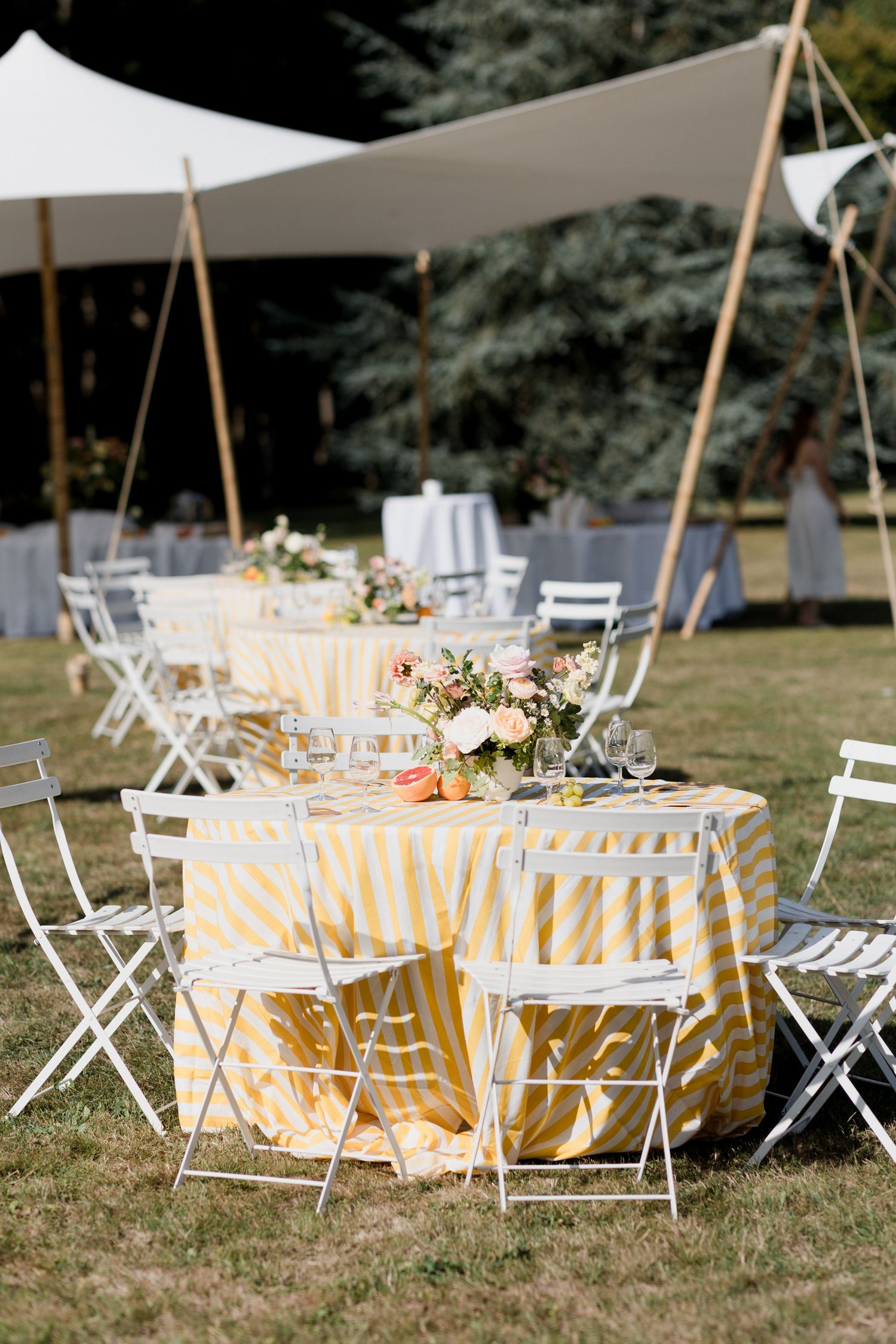Elegant outdoor wedding reception setup with yellow tablecloths, white folding chairs and floral centerpieces under a tent canopy.