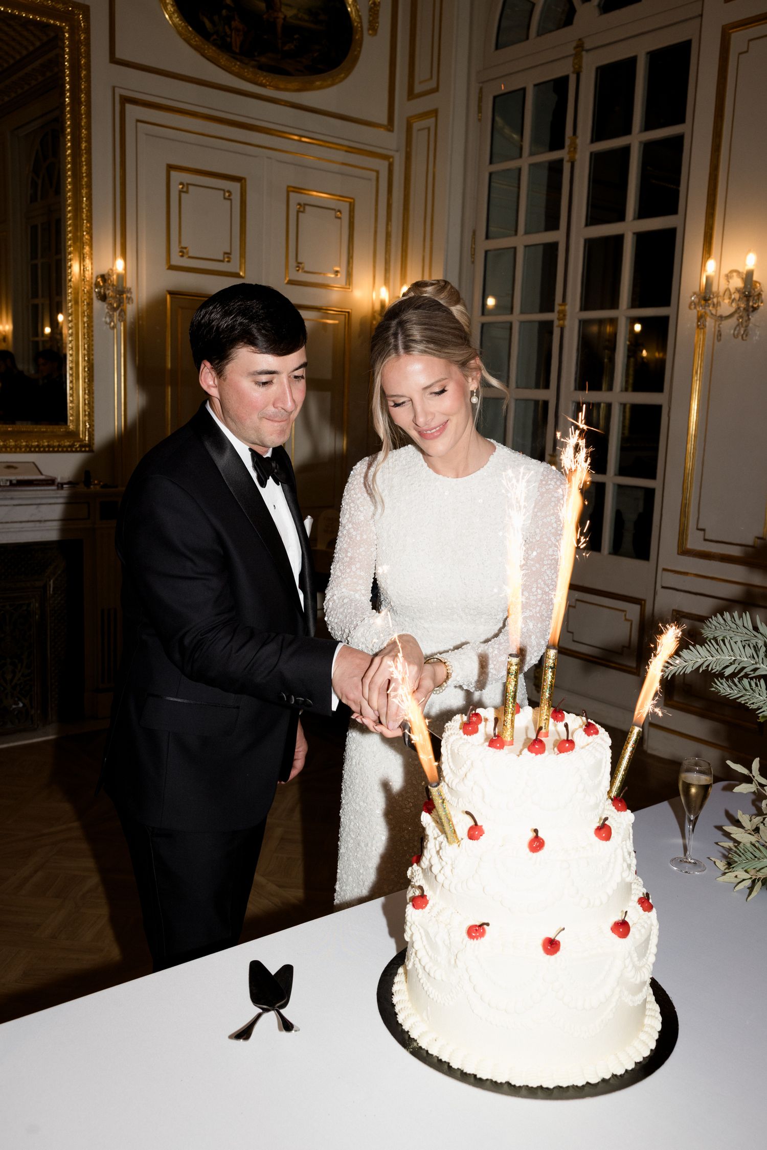 A couple cuts their elegant white wedding cake decorated with red accents in an ornate room with gold trim and candles.