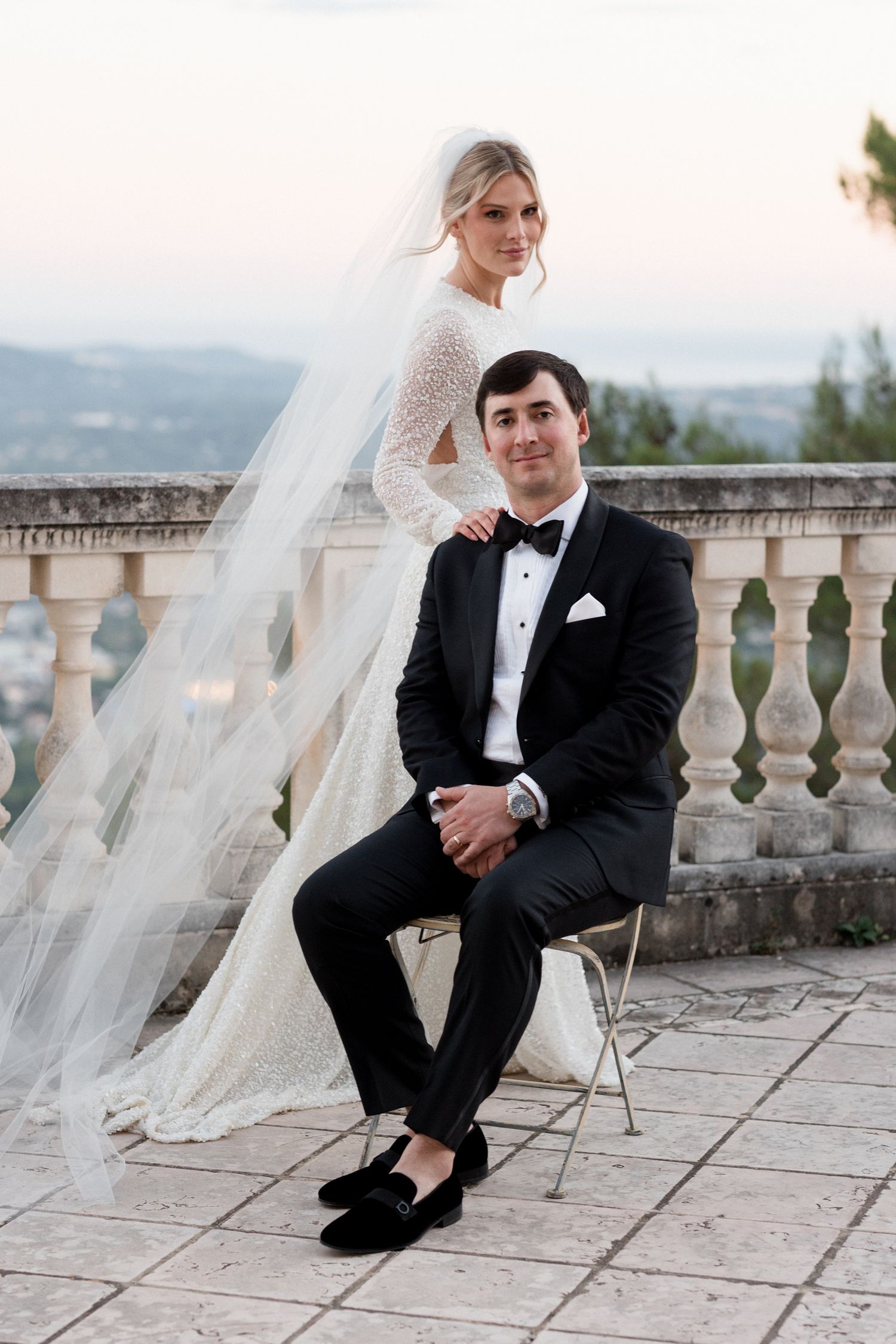 A bride and groom sit together on a stone balcony terrace overlooking scenic mountain views during their wedding day.