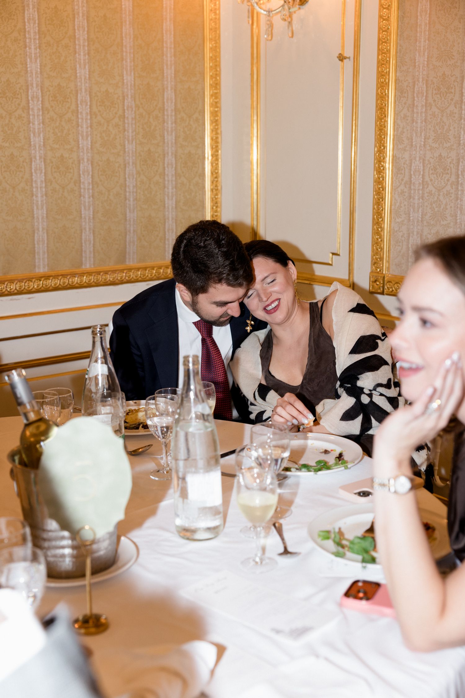 Elegant dinner party scene at a formal table with white tablecloth and ornate gold-trimmed walls in the background.