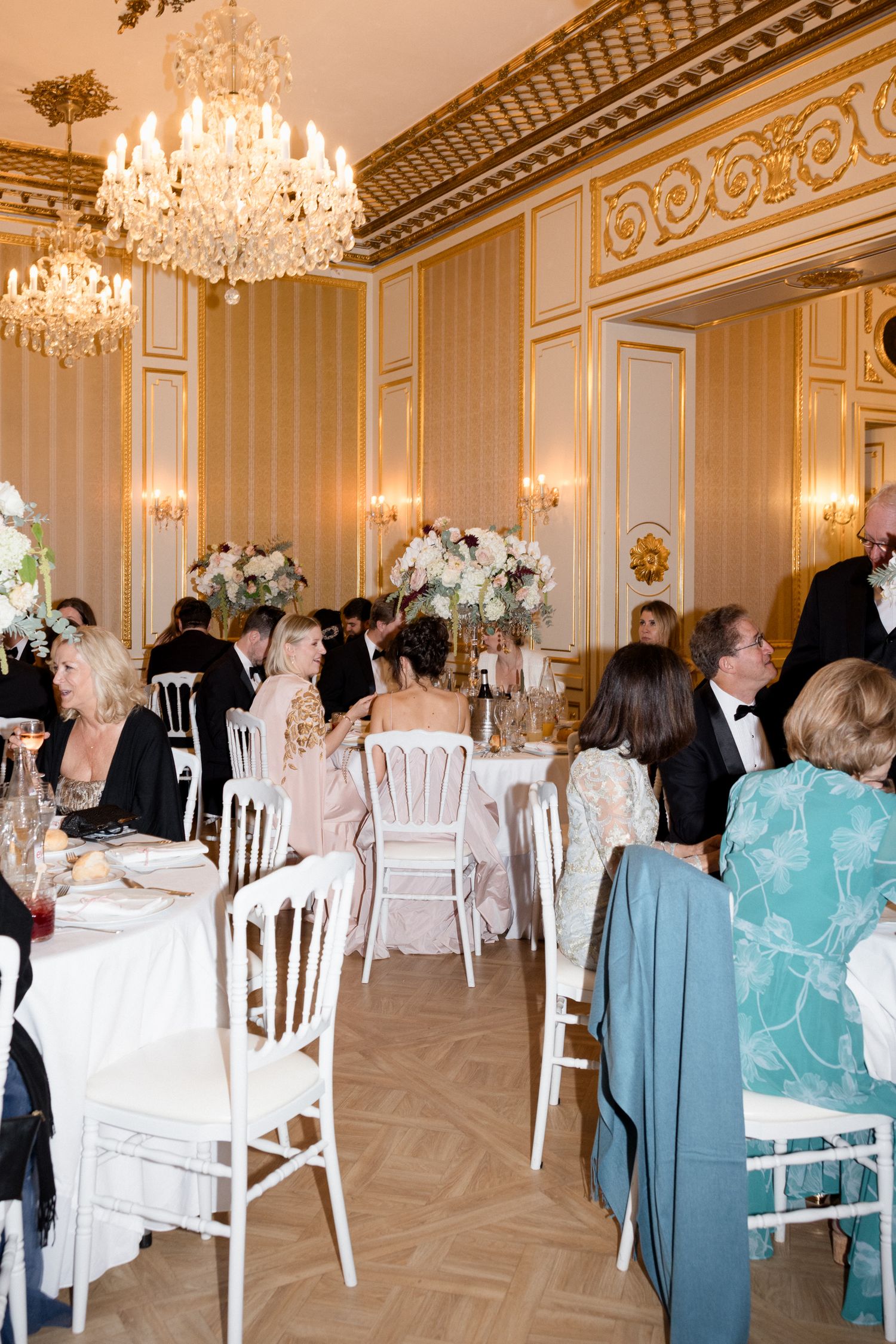 Elegant ballroom with crystal chandeliers, white chairs and tables set for a formal event with guests in formal attire.