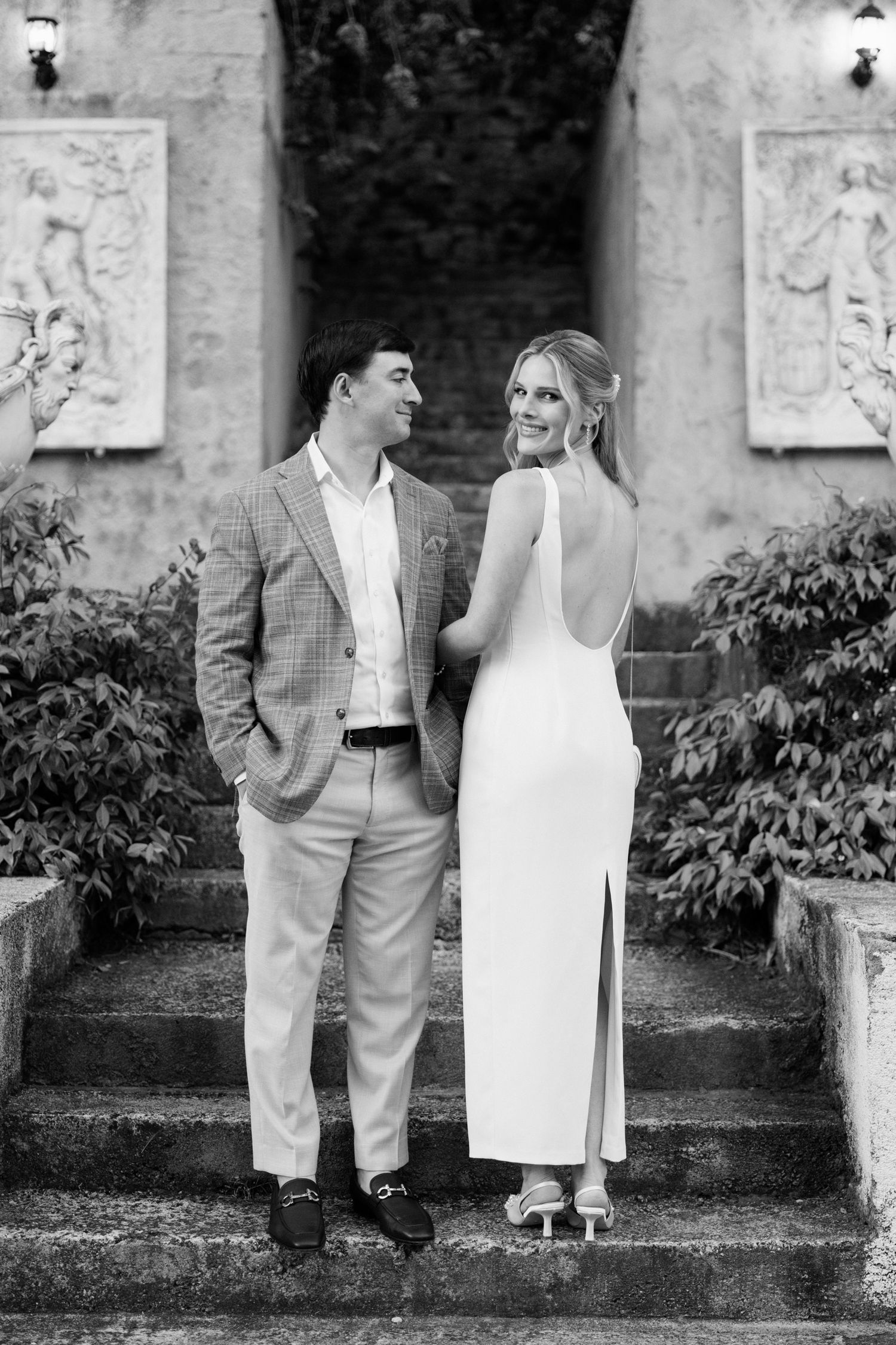A black and white wedding photo of a couple standing on stone steps with decorative wall art in an elegant garden setting.