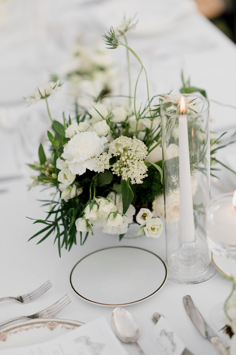 Elegant white wedding reception table setting with delicate floral arrangements, candles and minimalist place settings.
