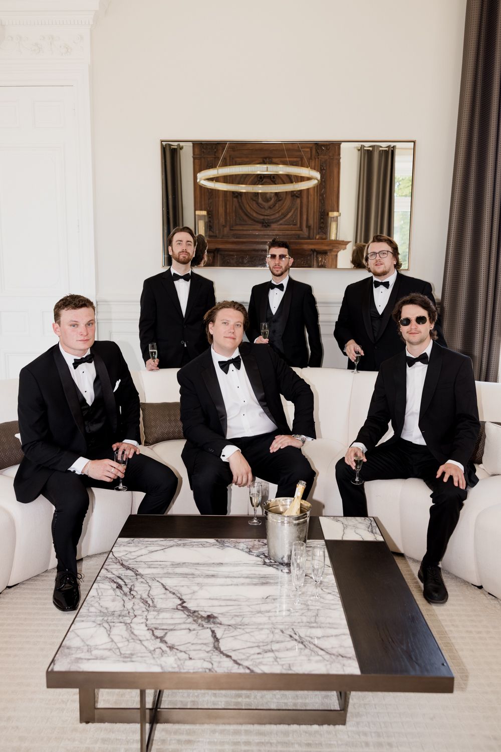 A group of groomsmen in black tuxedos sitting casually on a white couch in an elegant room with marble tables.