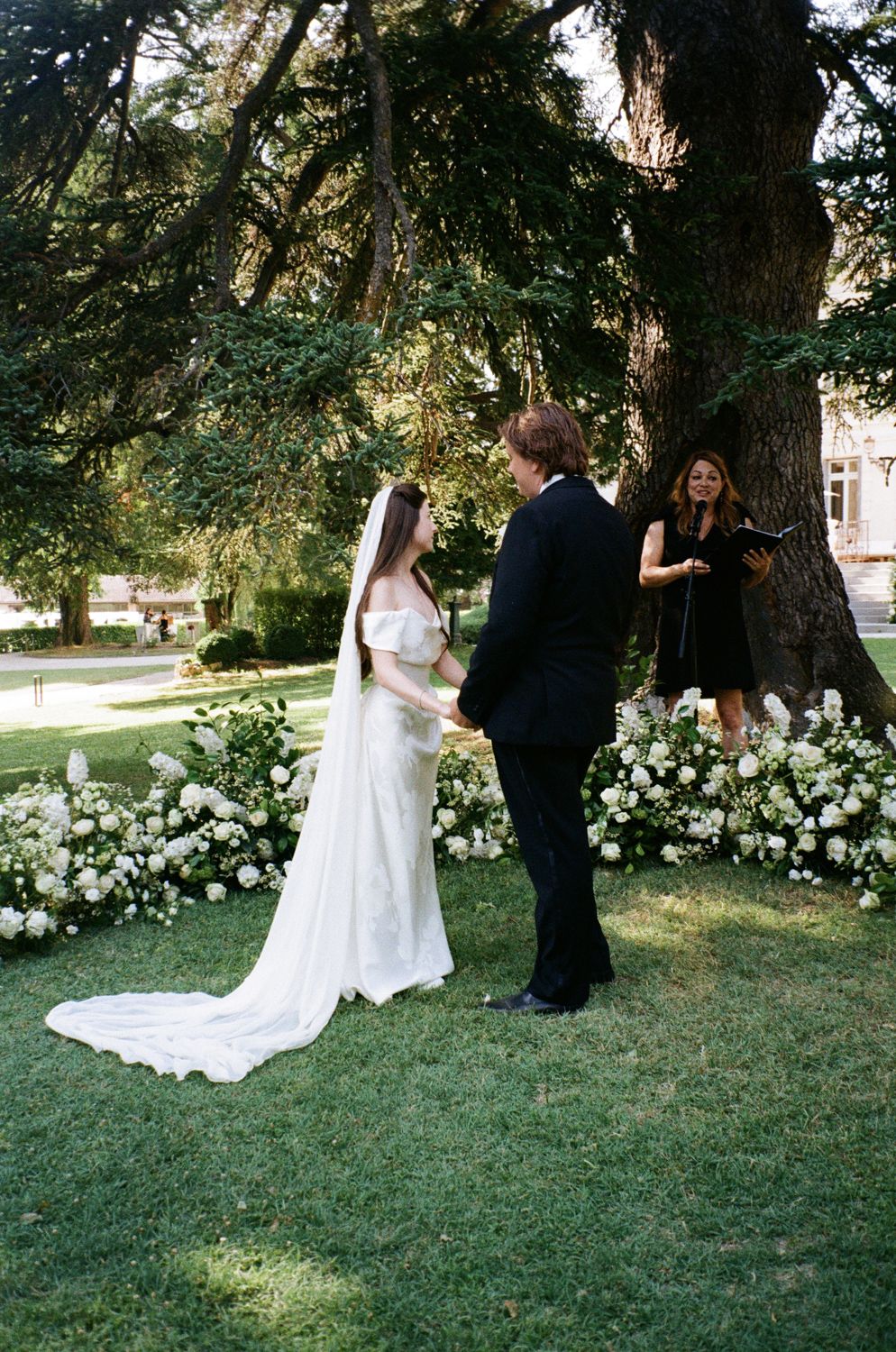 An outdoor wedding ceremony takes place under tall trees with white flowers blooming in the garden. Wedding on film, film photography