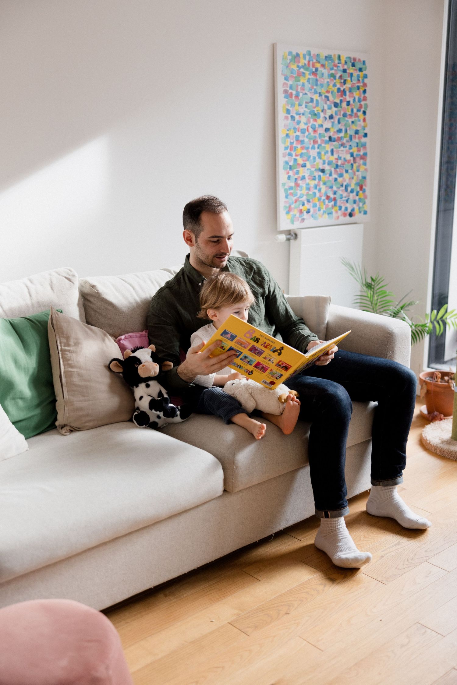 Father reads a colorful picture book with his toddler on a light gray sofa in a bright living room.