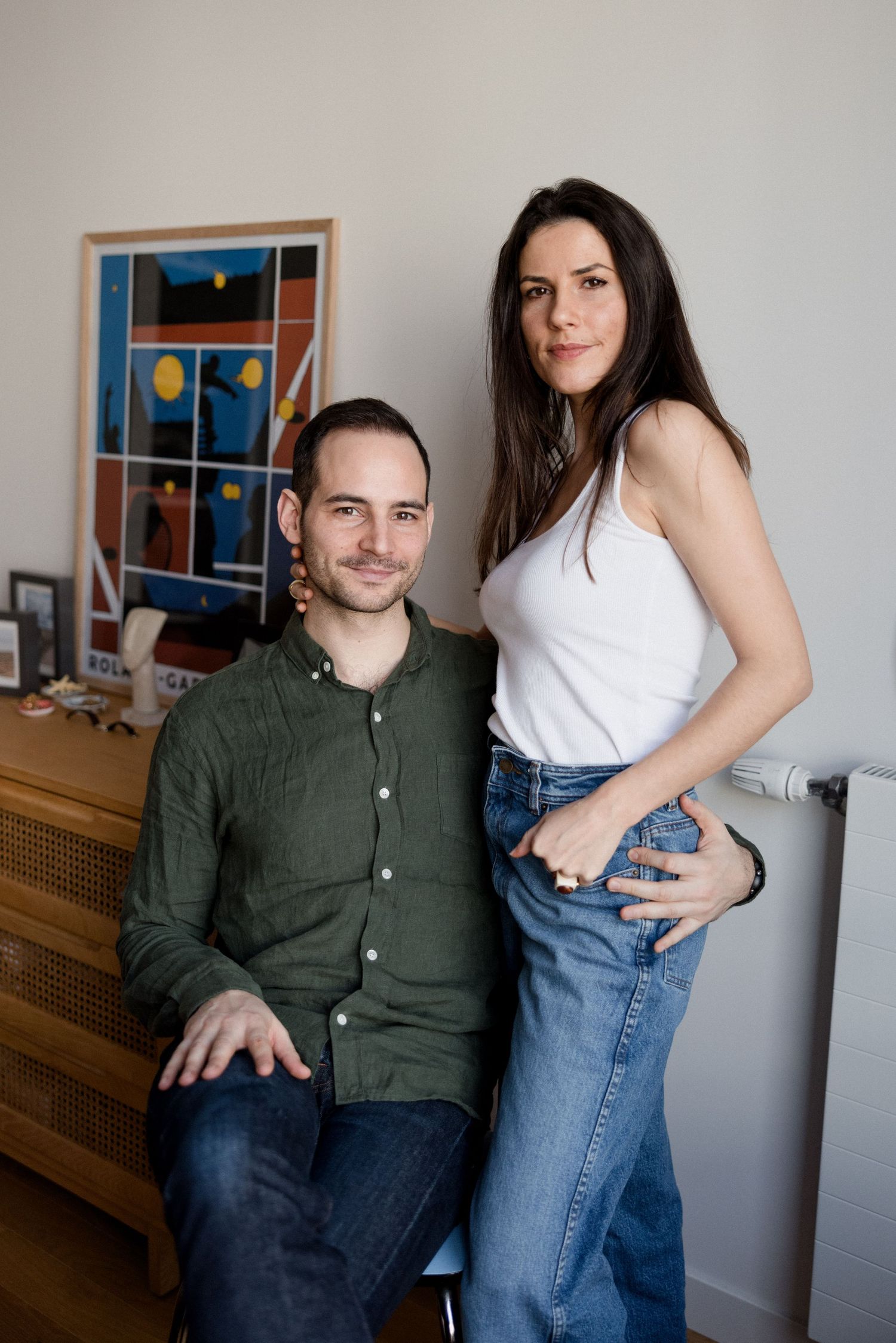 Man and woman pose together indoors in front of colorful artwork and wooden dresser.
