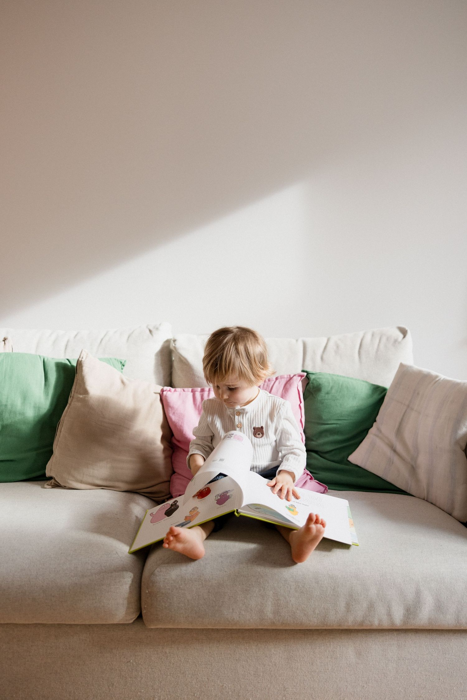 Toddler sitting barefoot on a beige sofa reading a colorful picture book surrounded by green and pink cushions.