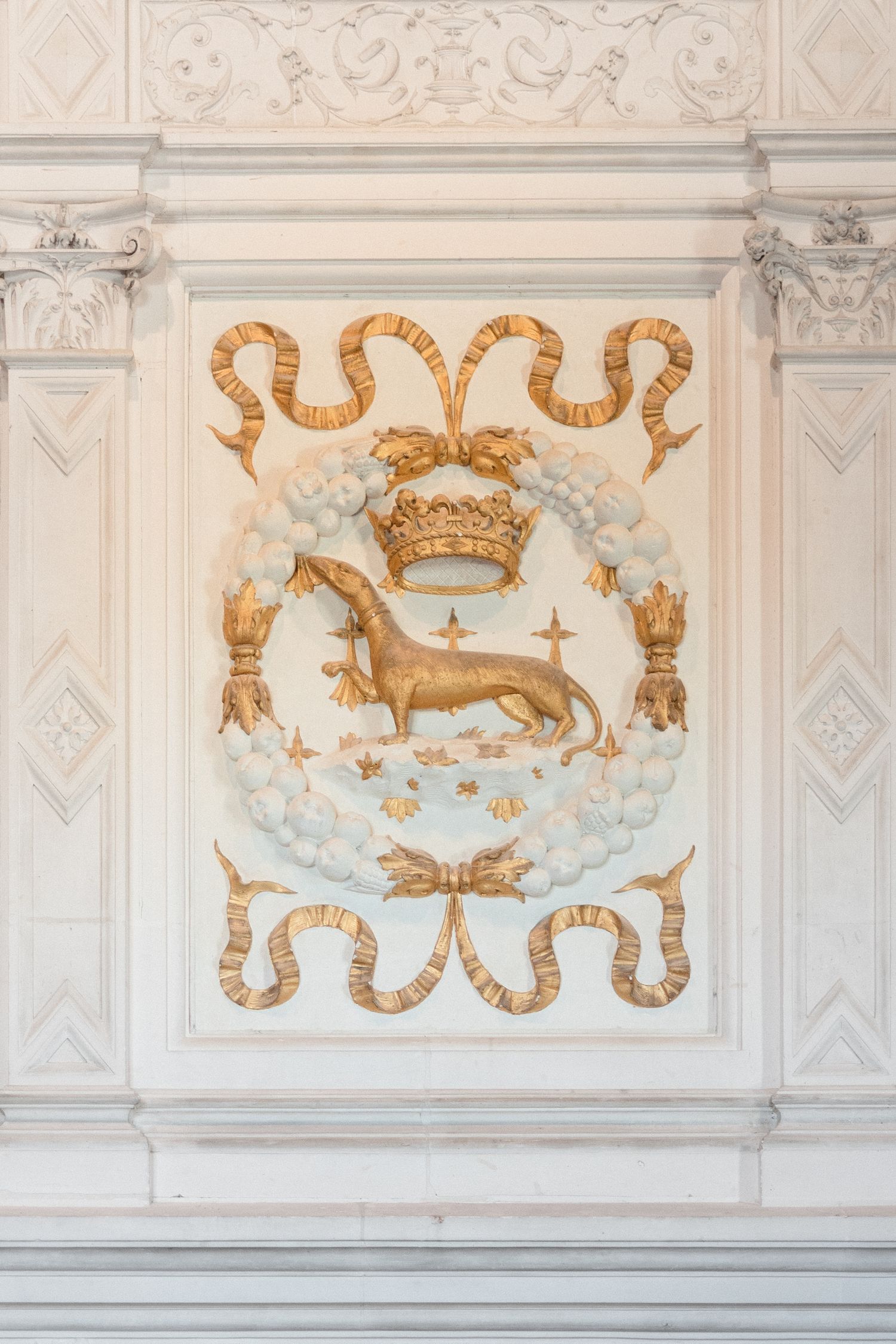 Golden heraldic relief featuring a dog beneath a crown, framed by serpents and floral motifs on an ornate white wall.