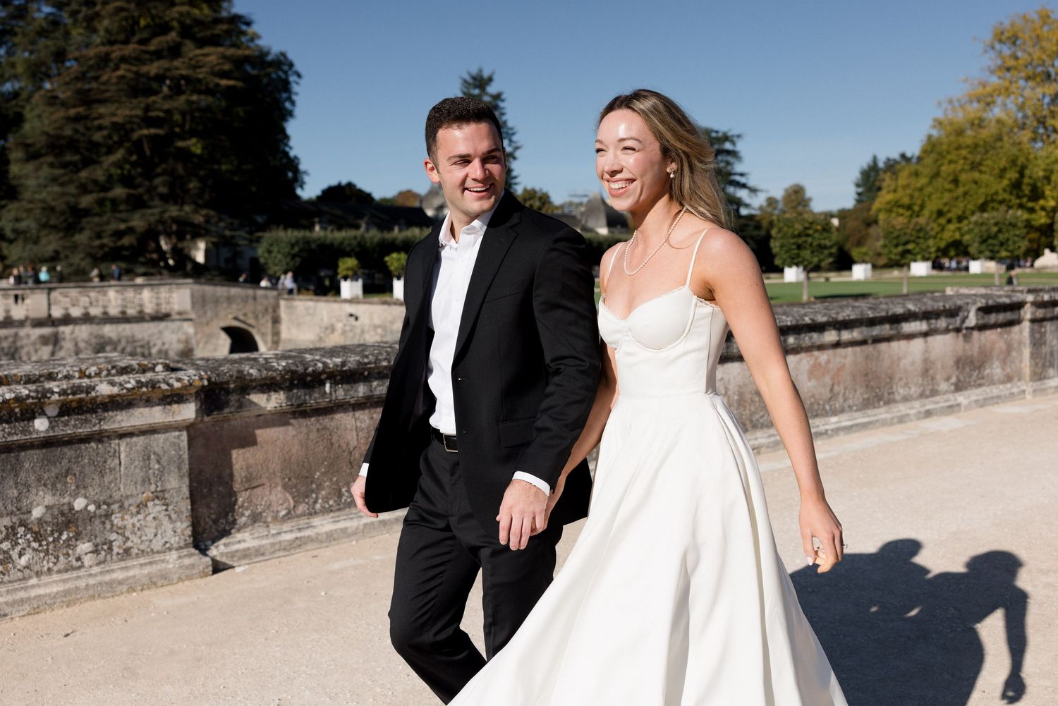 Smiling bride and groom in black tuxedo walking together in formal French chateau garden.