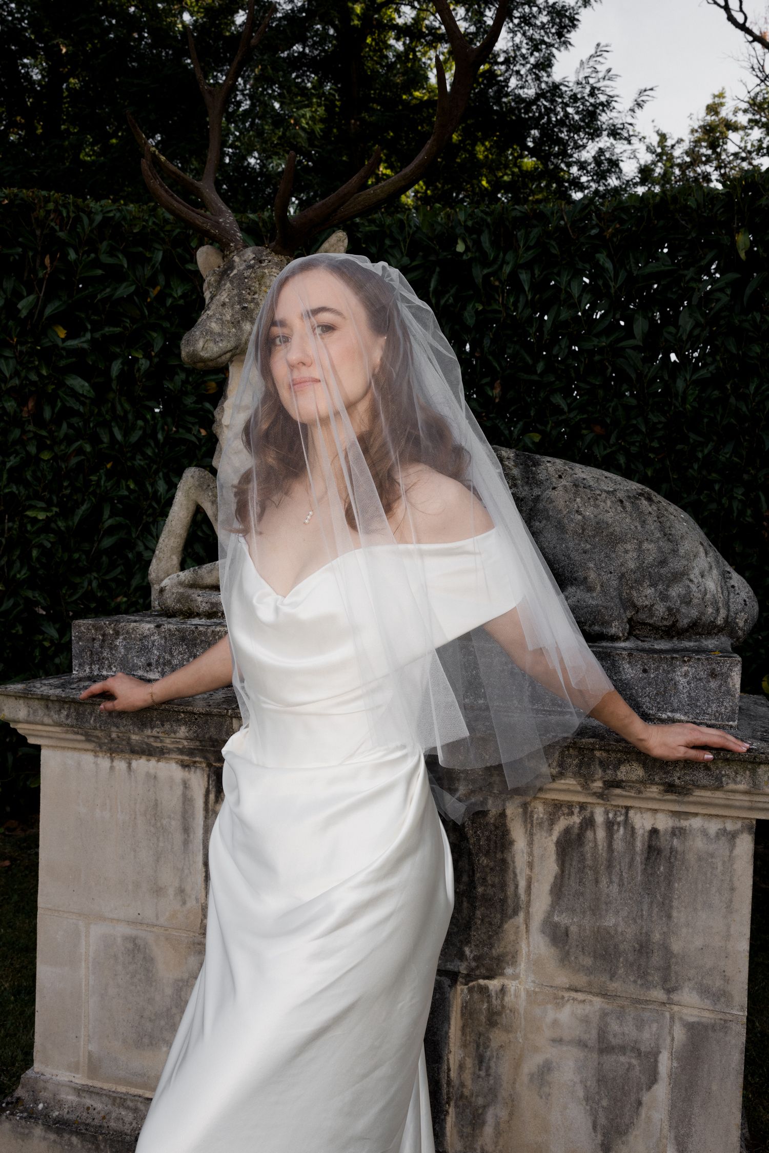 A person in a flowing white wedding dress poses dramatically against stone architecture in an outdoor garden setting. Paris edito Tours