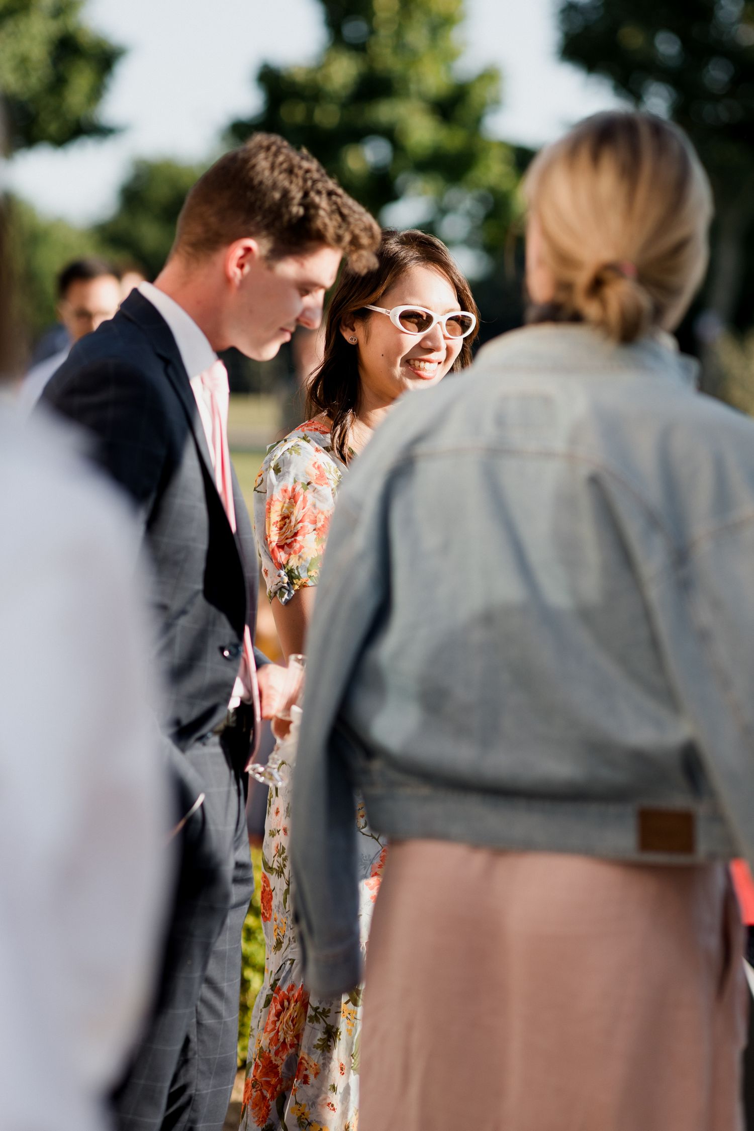 A sunny outdoor gathering shows people in formal attire including a floral dress and dark suit interacting at a social event.