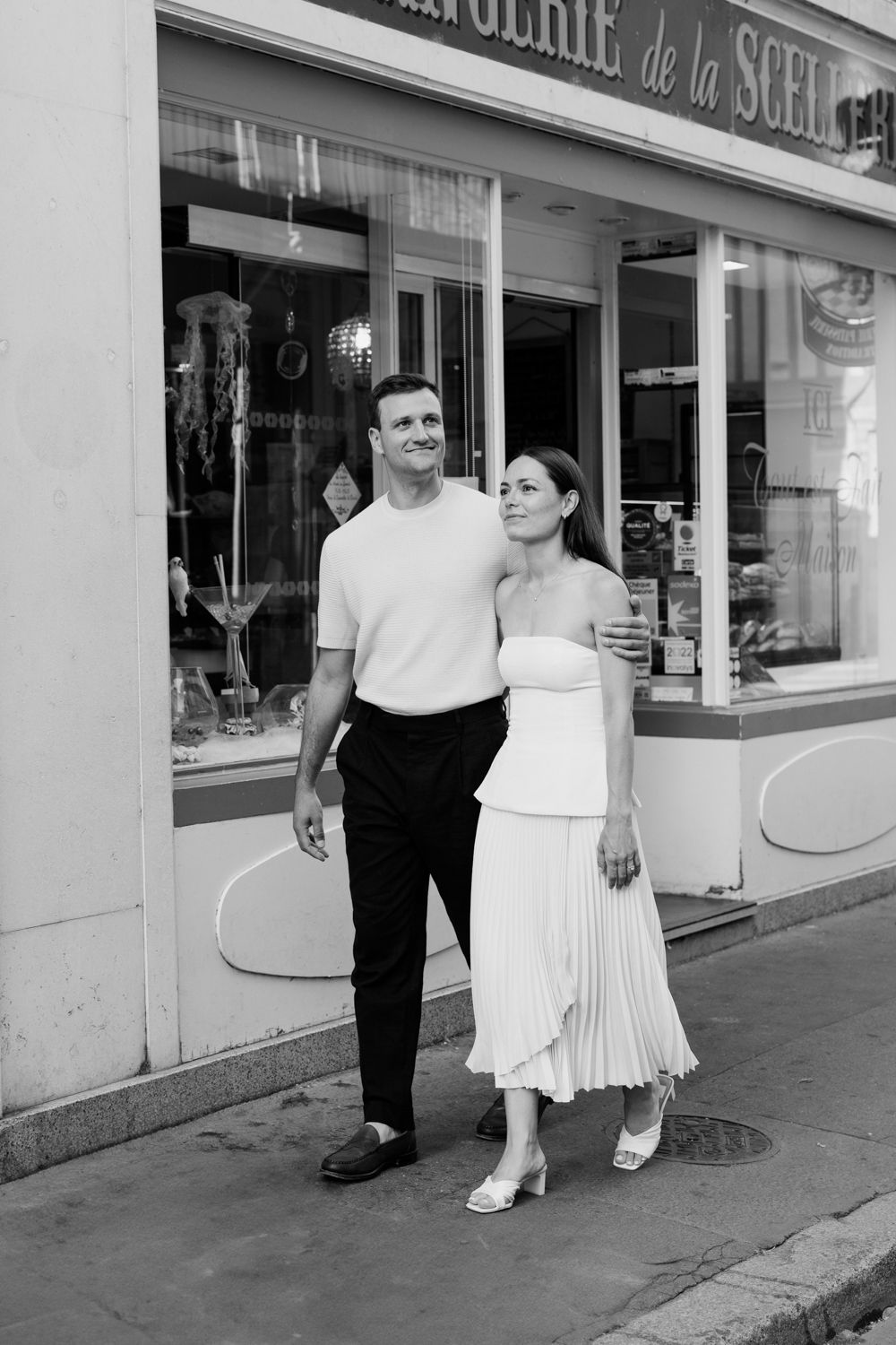 A black and white photo showing two people walking past storefronts with French signage on a city street.