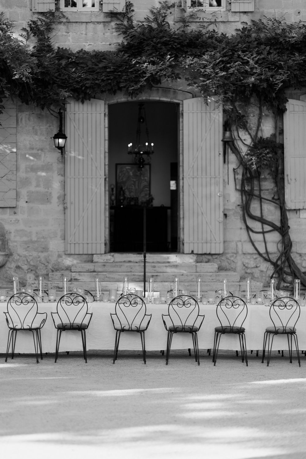 A row of six empty vintage wrought iron chairs lined up against a stone wall with climbing vines and an open doorway.