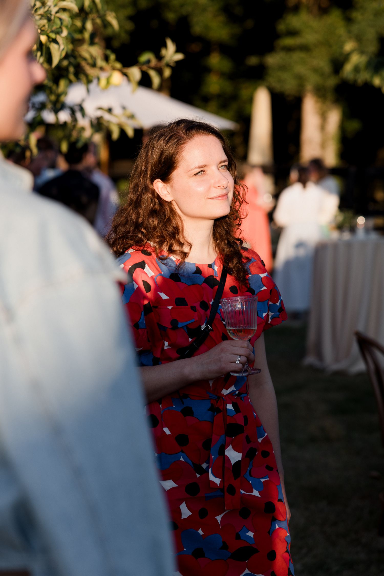 A person in a red patterned dress holds a wine glass at an outdoor evening event.