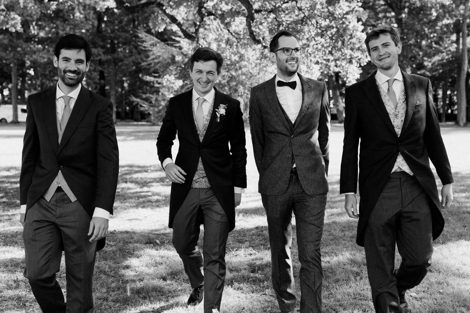 A group of groomsmen in dark suits walk together outdoors while smiling and laughing during a wedding photoshoot.