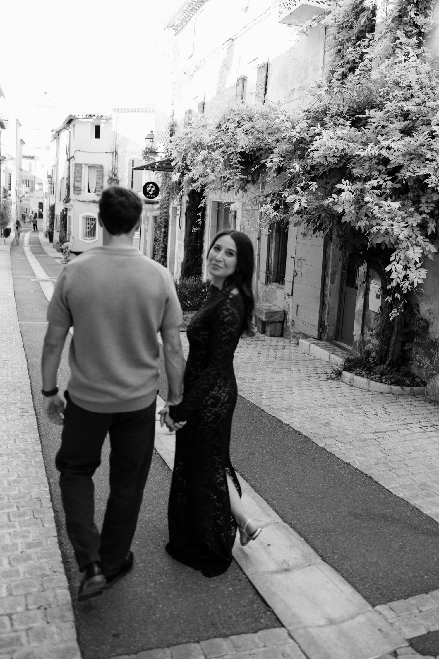 Two people walking down a charming cobblestone street lined with trees and buildings in black and white. Saint Remy de Provence
