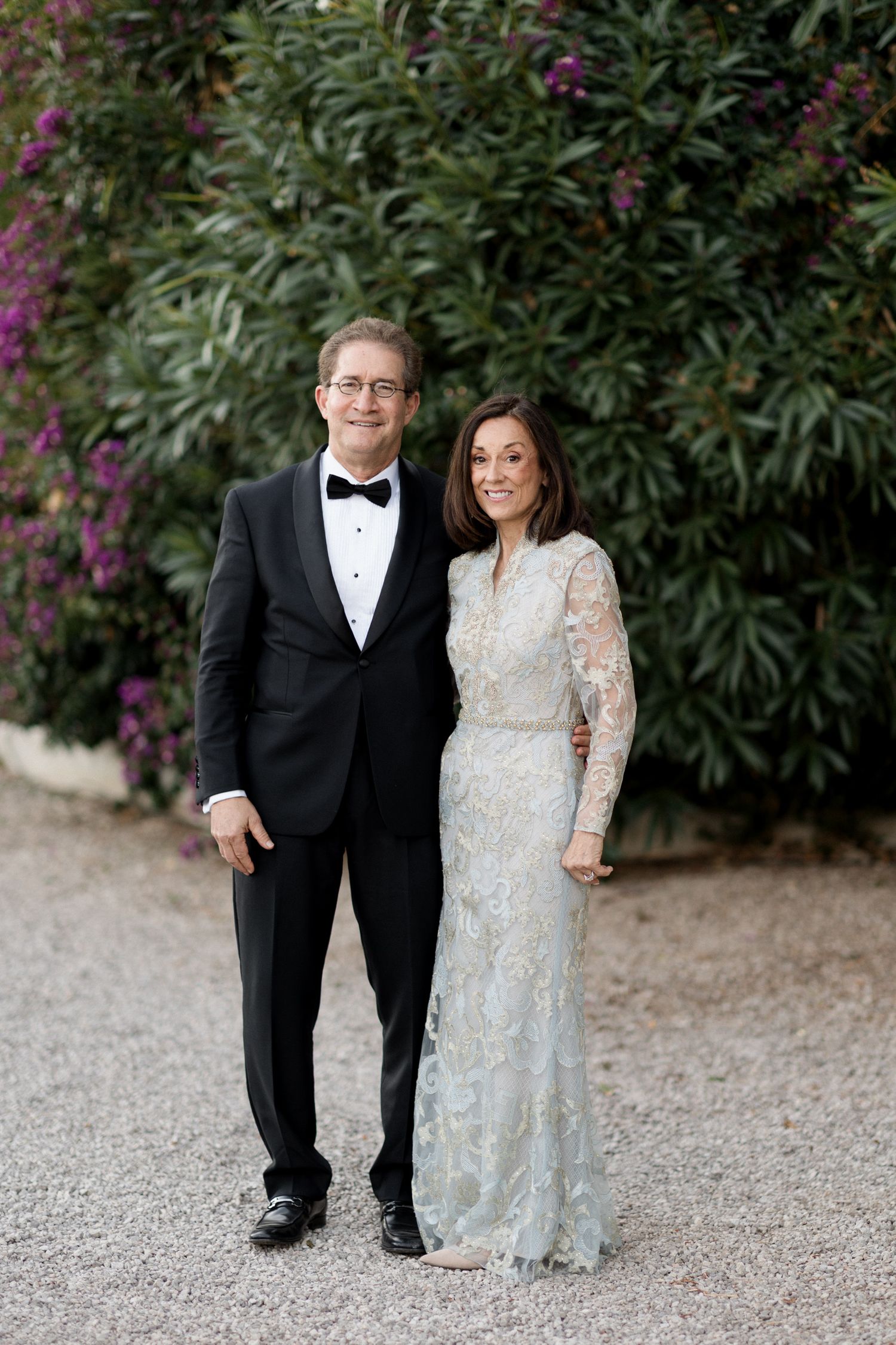 A couple in formal attire poses together on a gravel path with flowering bushes in the background.