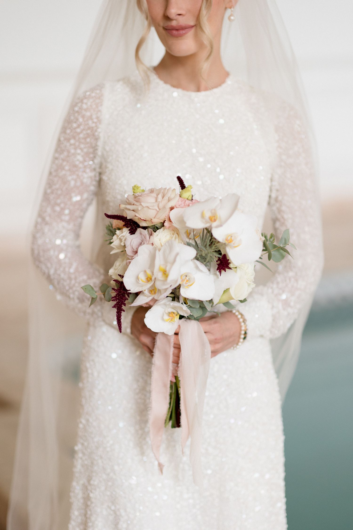 Bride in long sleeve sequined wedding dress holds elegant white and blush floral bouquet with trailing ribbons. Mariage Grasse, France