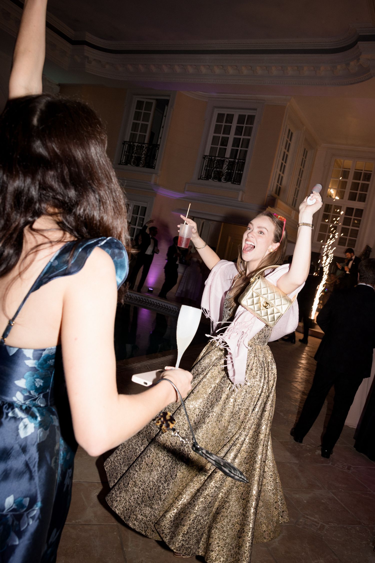 People dancing and celebrating at an evening party in a ballroom with gold dresses and formal attire.