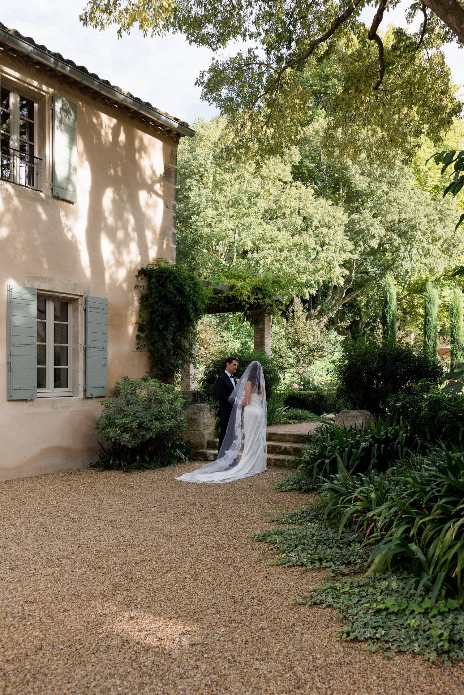 A romantic garden path leads to a charming French country house with pale walls and blue shutters in dappled sunlight.