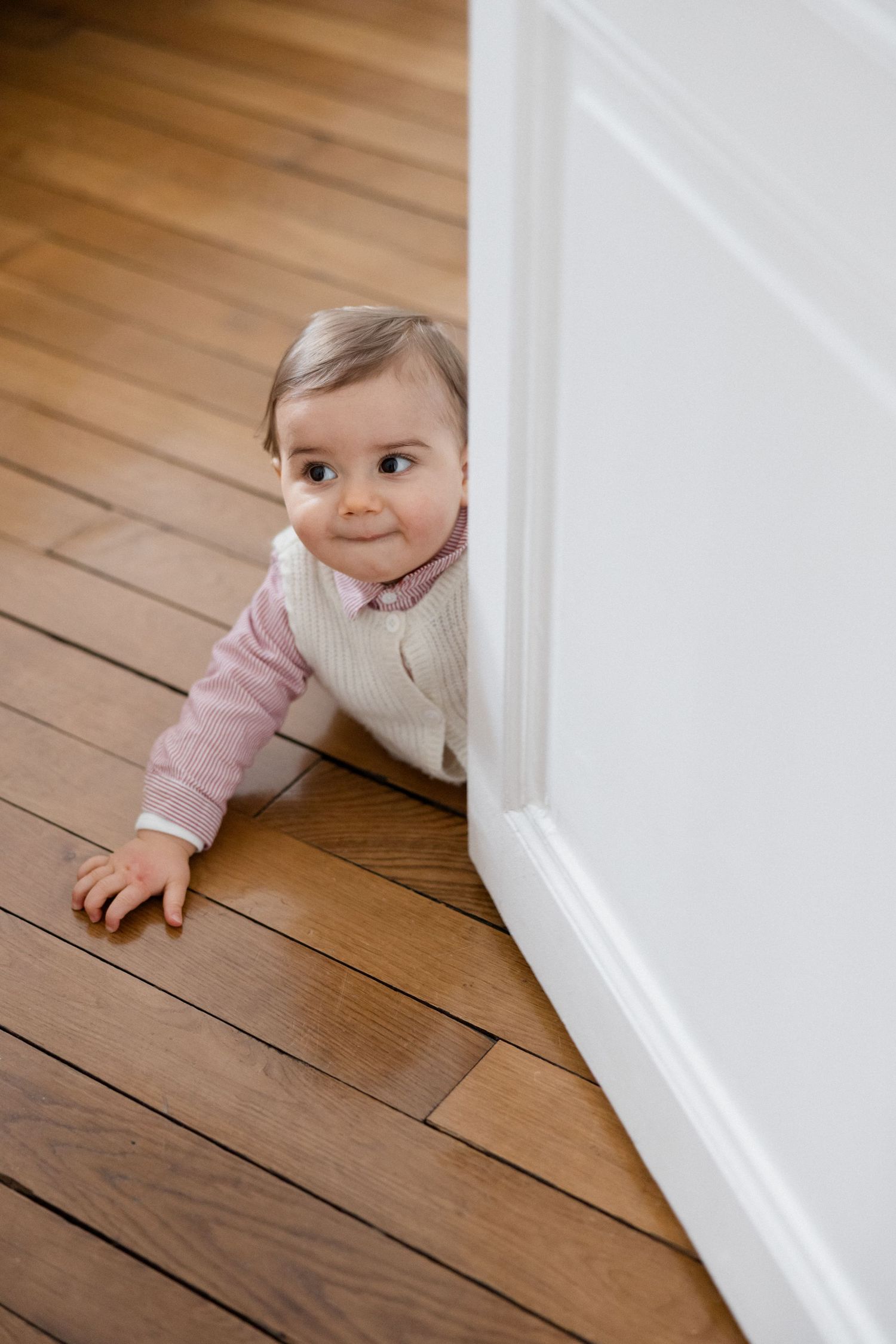 Curious baby in pink outfit peeking around a white wall corner on warm hardwood floors.