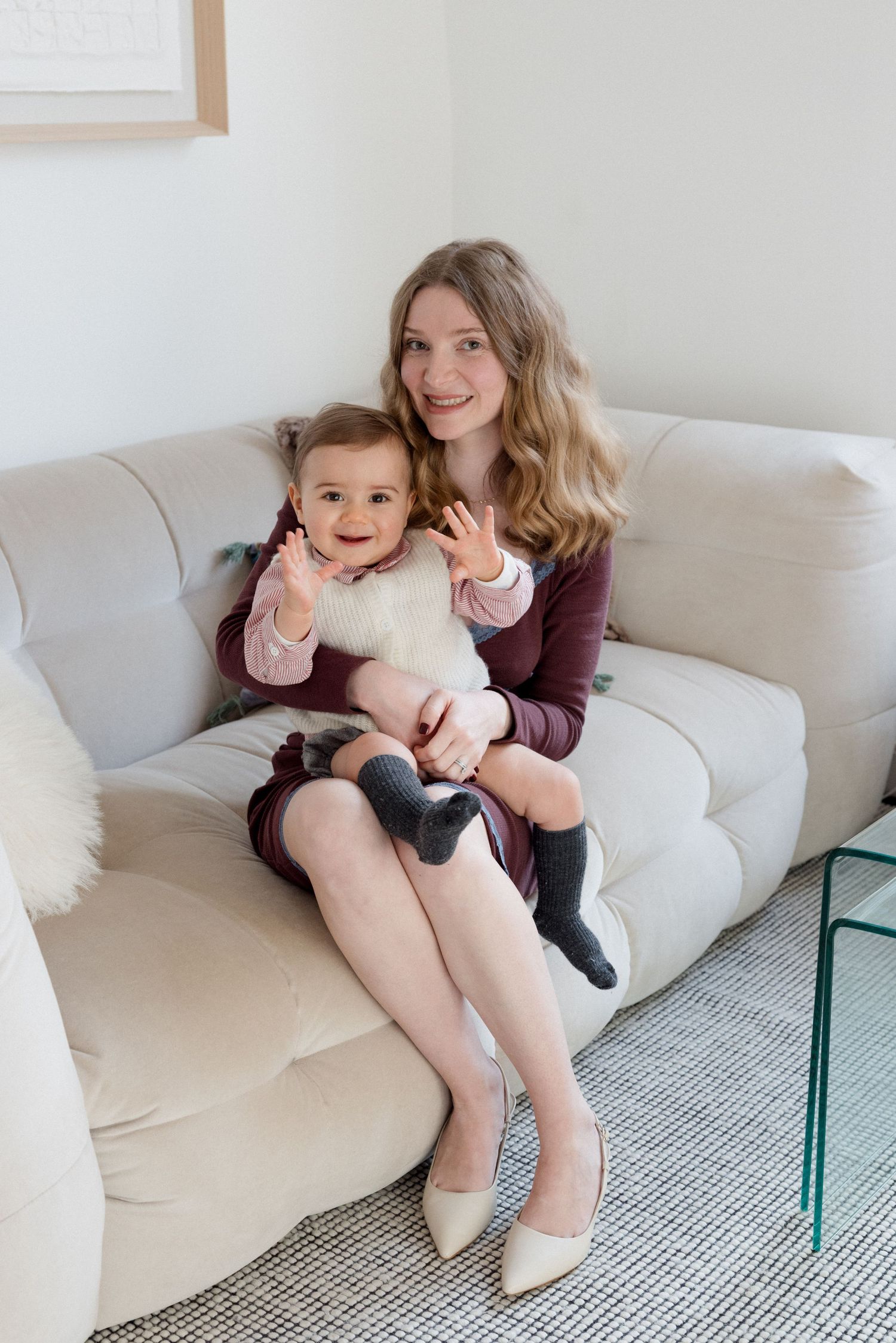 A smiling young woman in a burgundy dress sits on a cream sofa holding a happy baby waving at the camera.