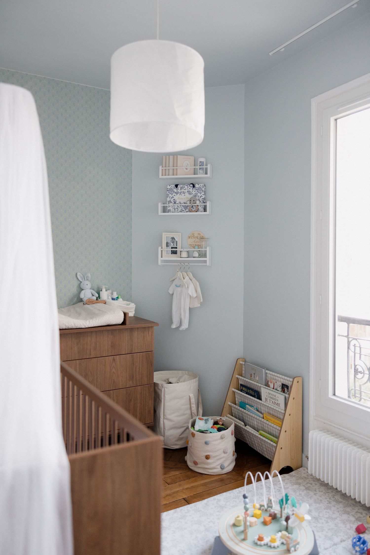 Soft blue nursery with wooden crib, dresser, toy bookshelf, and white pendant light on hardwood floors.