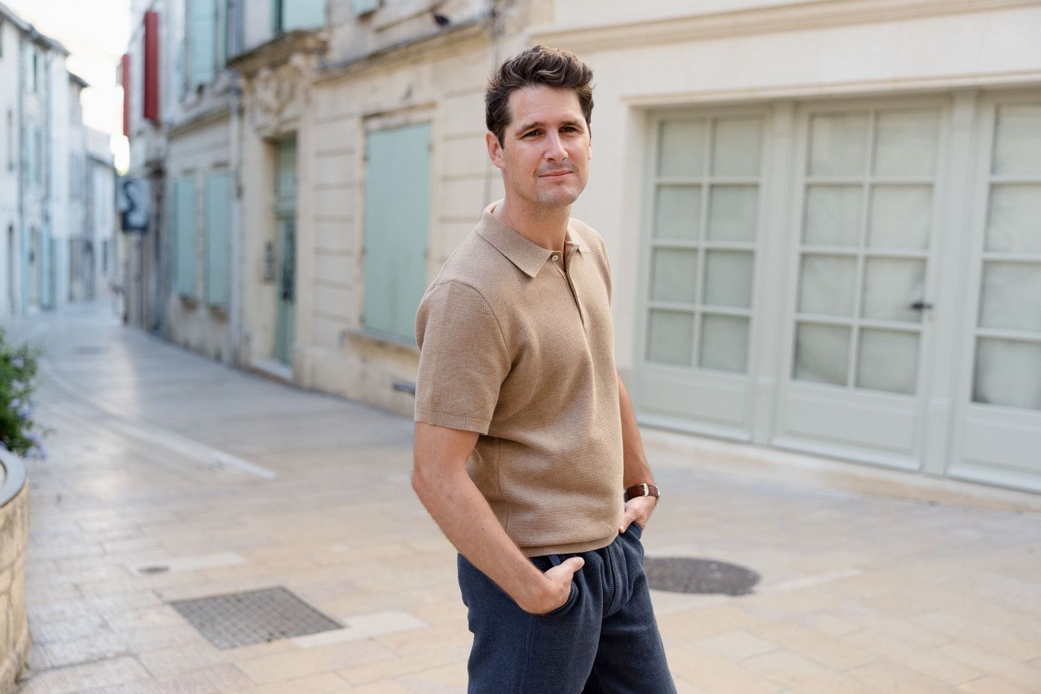 A person wearing a beige sweater and dark pants walks down a narrow European-style street with historic buildings.