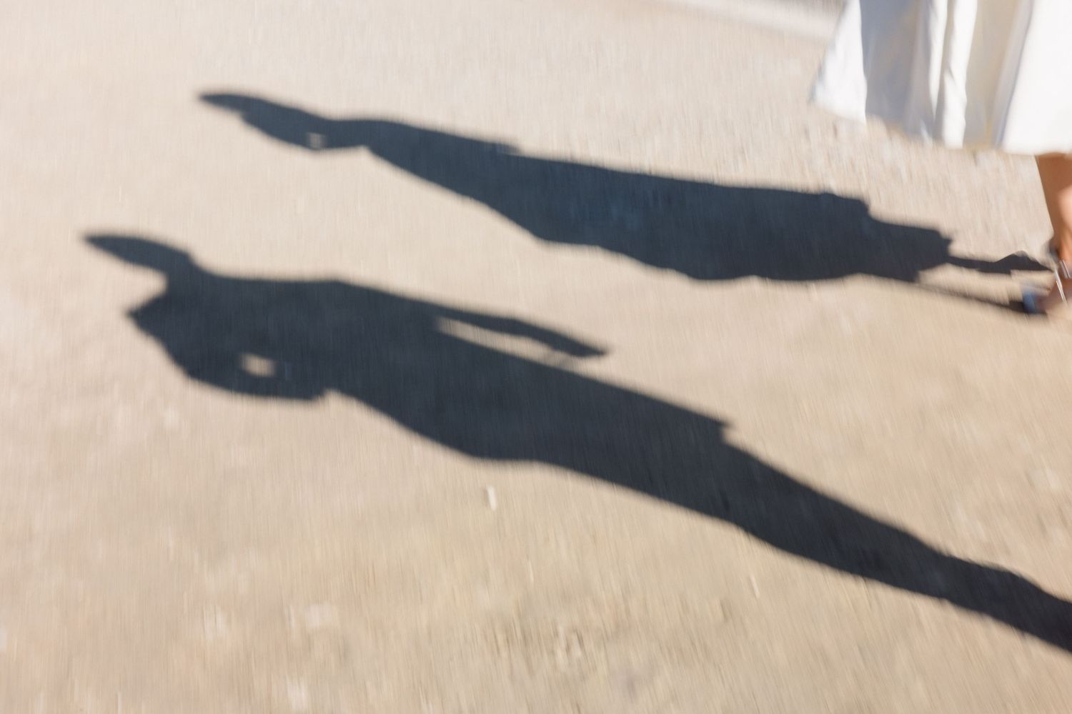 Two elongated shadows of people cast on sandy ground in bright sunlight, with a white dress visible at the edge.