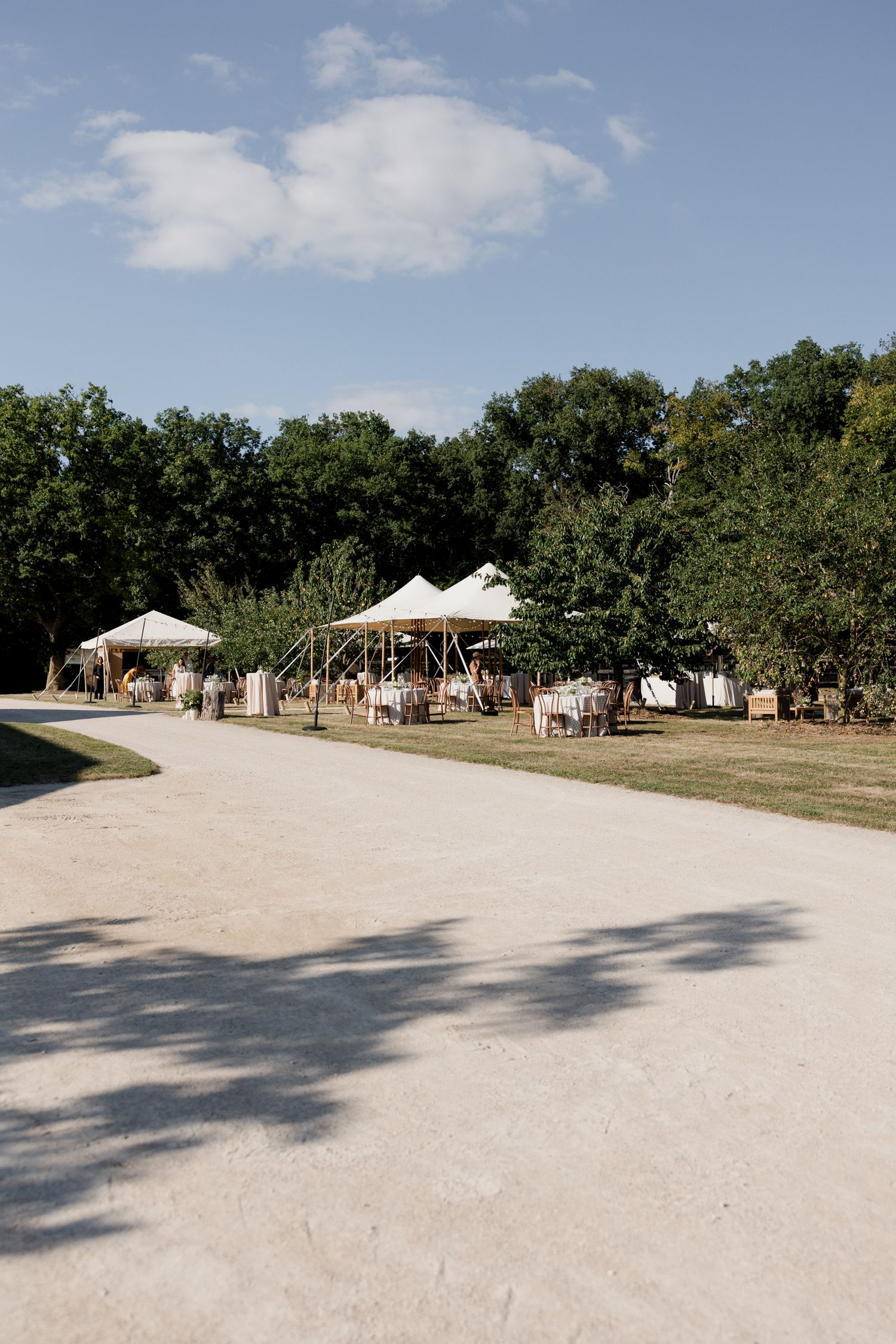 White tents and market stalls line a sandy path under green trees at an outdoor festival venue.