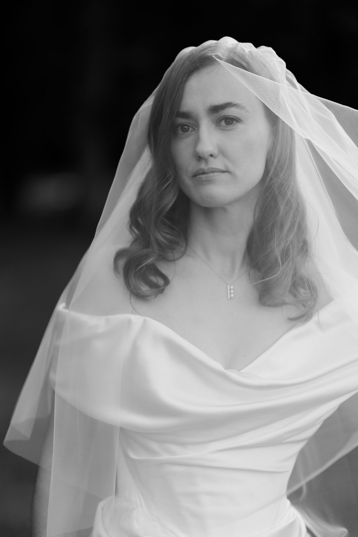Black and white portrait of a bride in an off-shoulder wedding dress with flowing veil and soft curled hair.