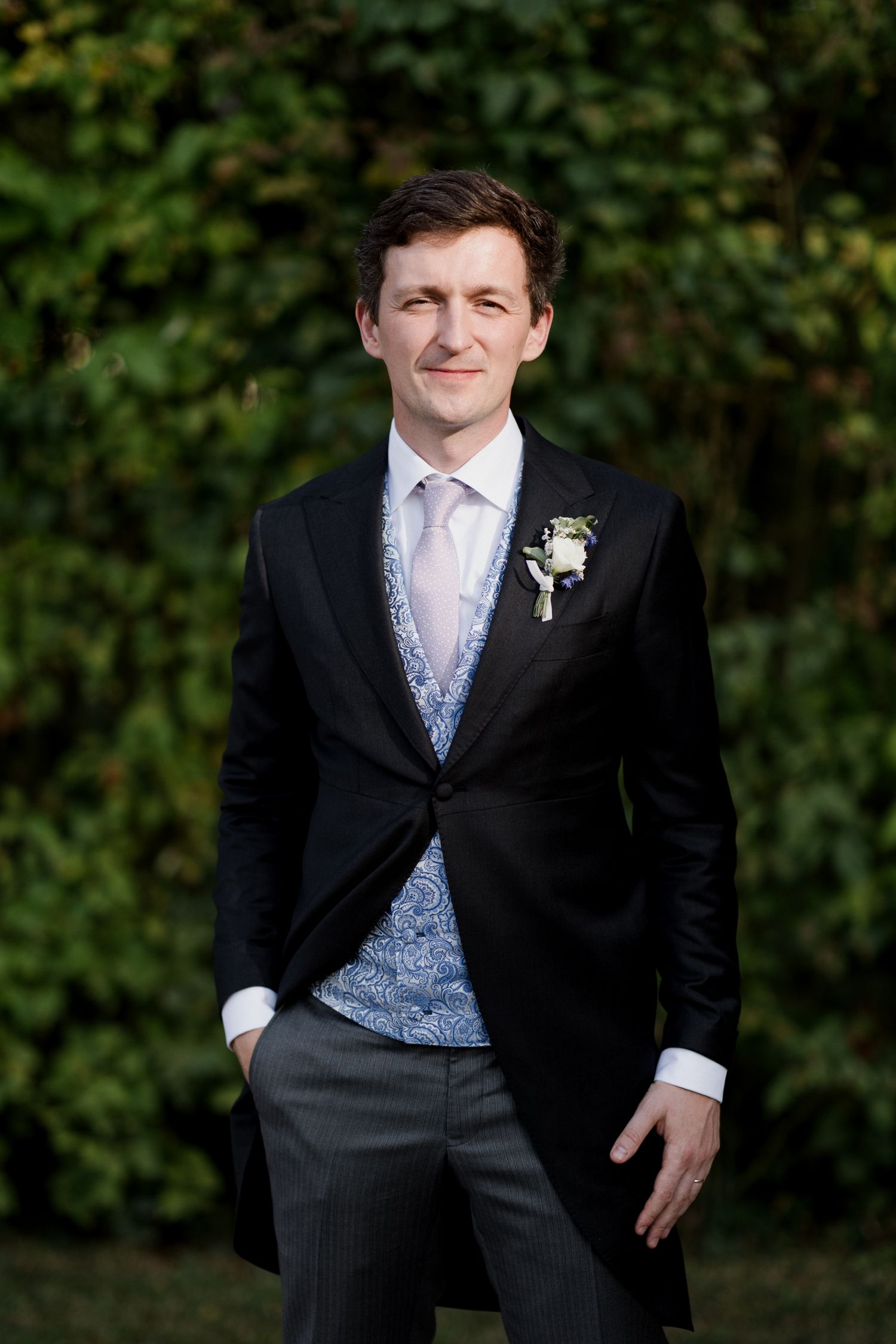 A groom in a formal black suit with blue vest and white tie poses against a green foliage backdrop.