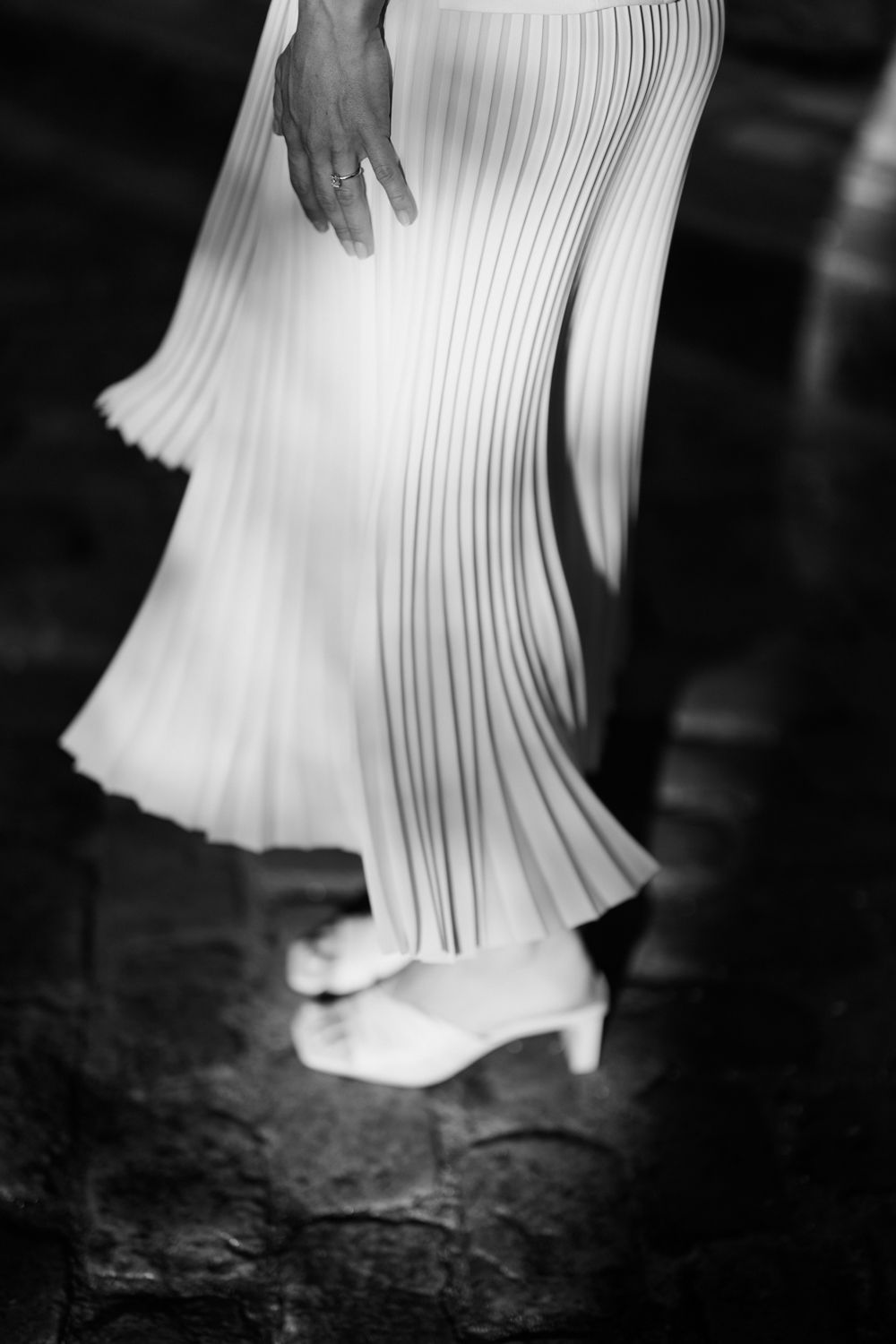 A black and white closeup of feet in white heels beneath a pleated white flowing dress skirt.