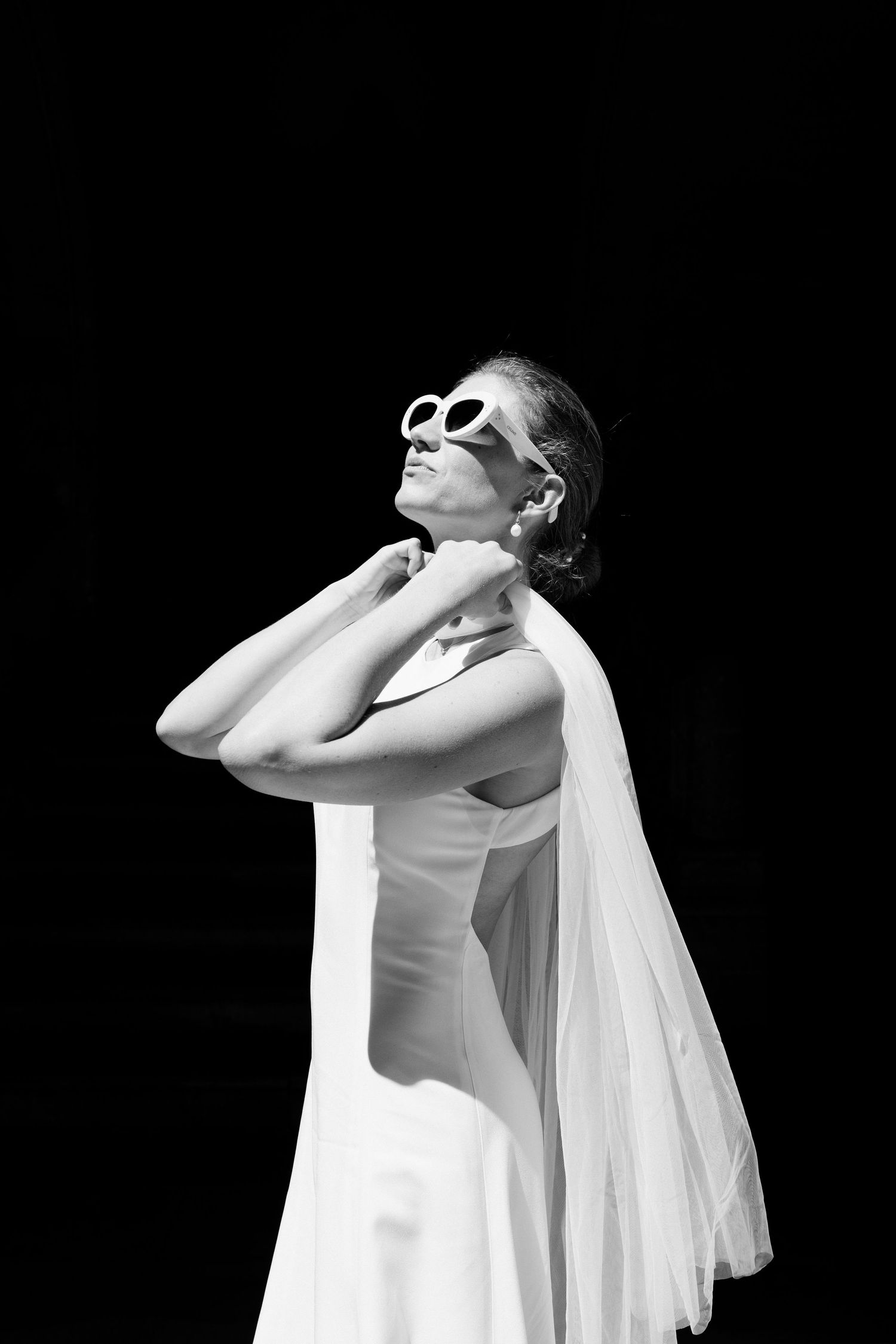Black and white photo of a bride in white gown and sunglasses, posing elegantly against a dark background.