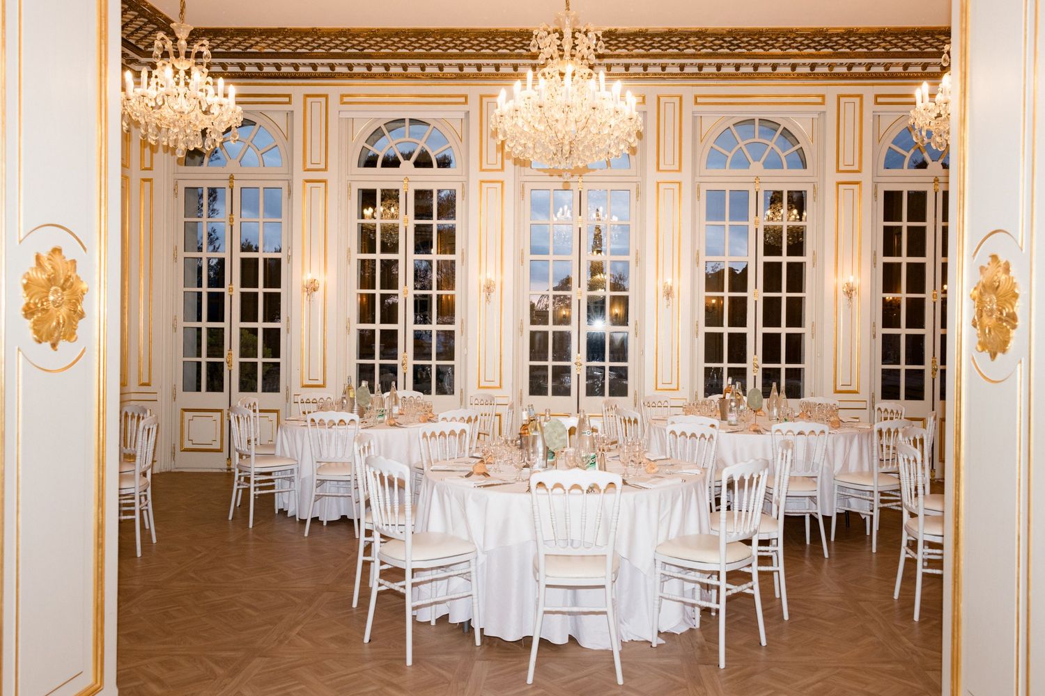 Panoramic view of an elegant white ballroom with crystal chandeliers, ornate wall sconces, and round tables set for dining. destination wedd