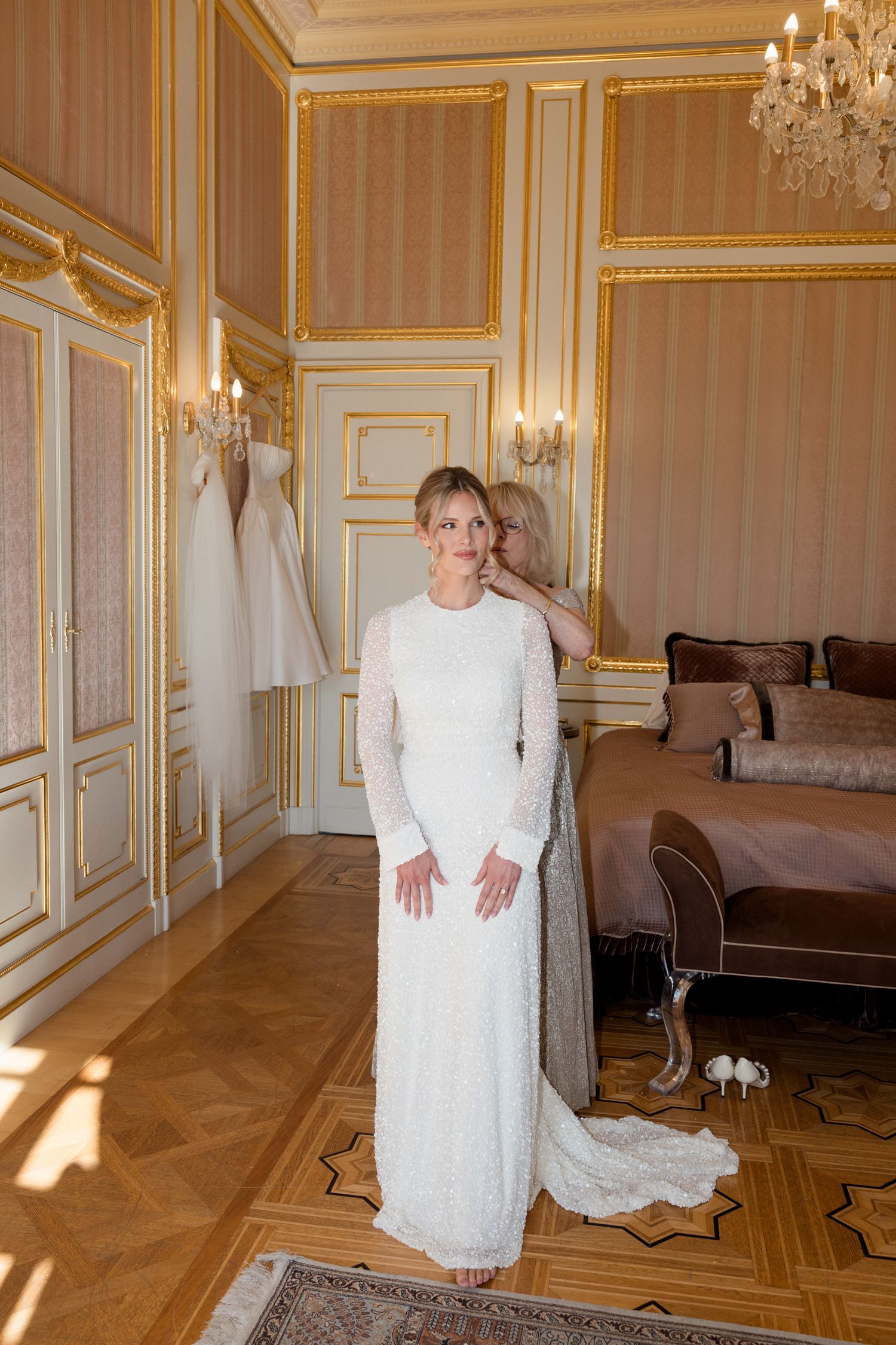 Elegant figure in white lace dress stands in ornate room with crystal chandeliers and herringbone wood floors.