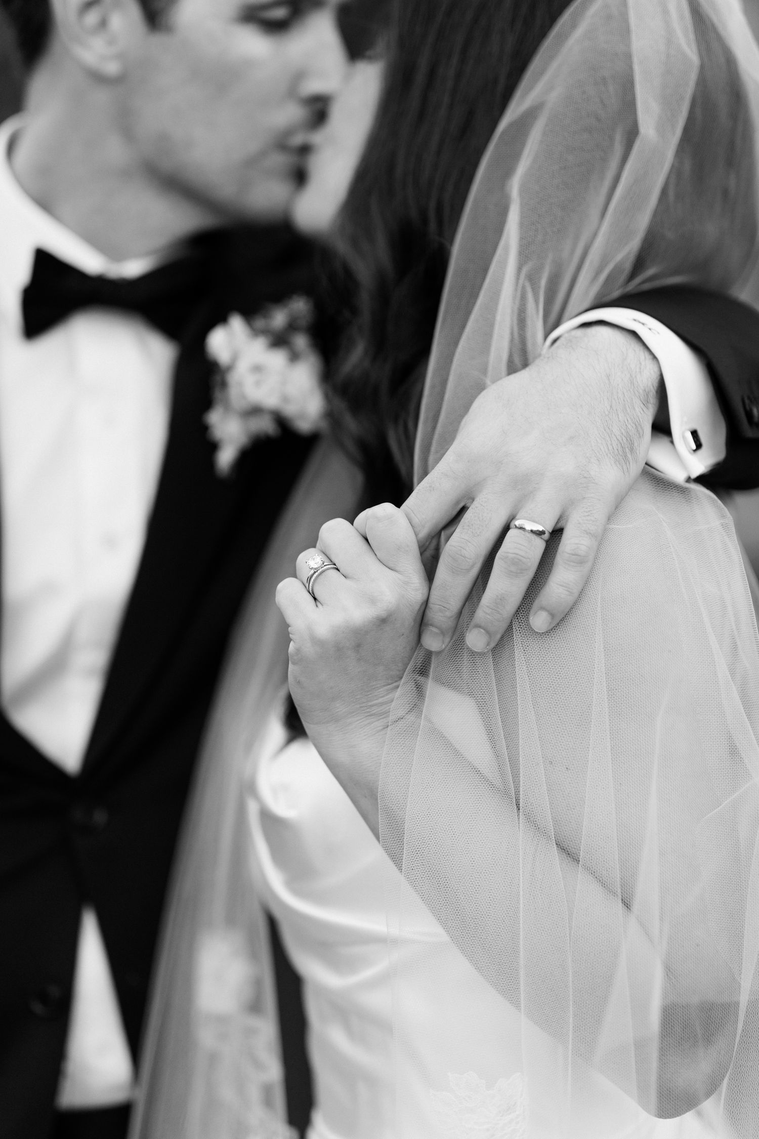 Black and white wedding photo showing hands with rings and a delicate veil against formal attire with boutonniere.